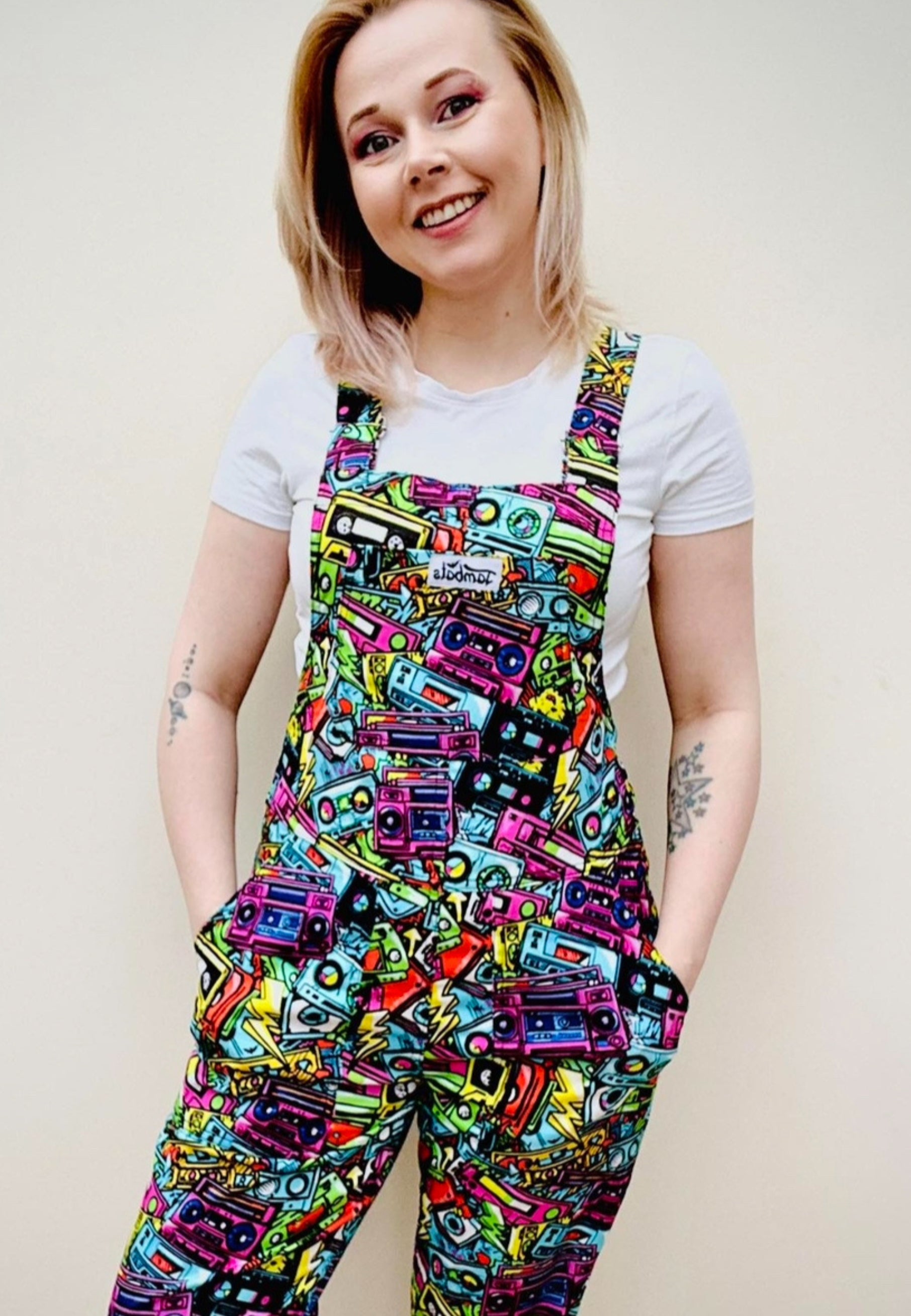 Woman wearing bright coloured dungarees with a retro 80s 90s print of graffiti street art music cassettes and ghetto blasters. Casual styling. 