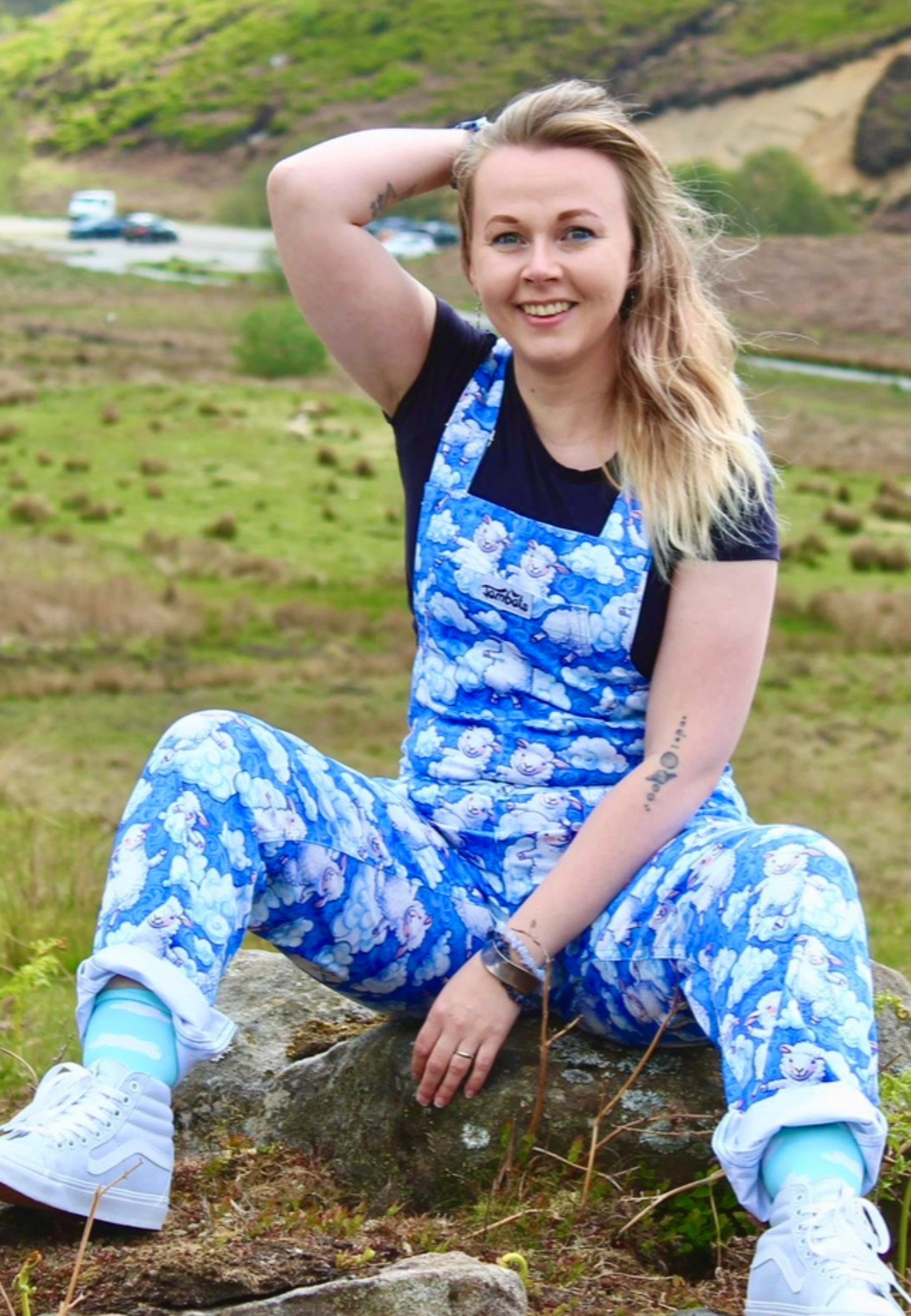 Model wearing a blue fluffy cloud and fluffy sheep patterned dungarees sitting on a rock in a grassy field.