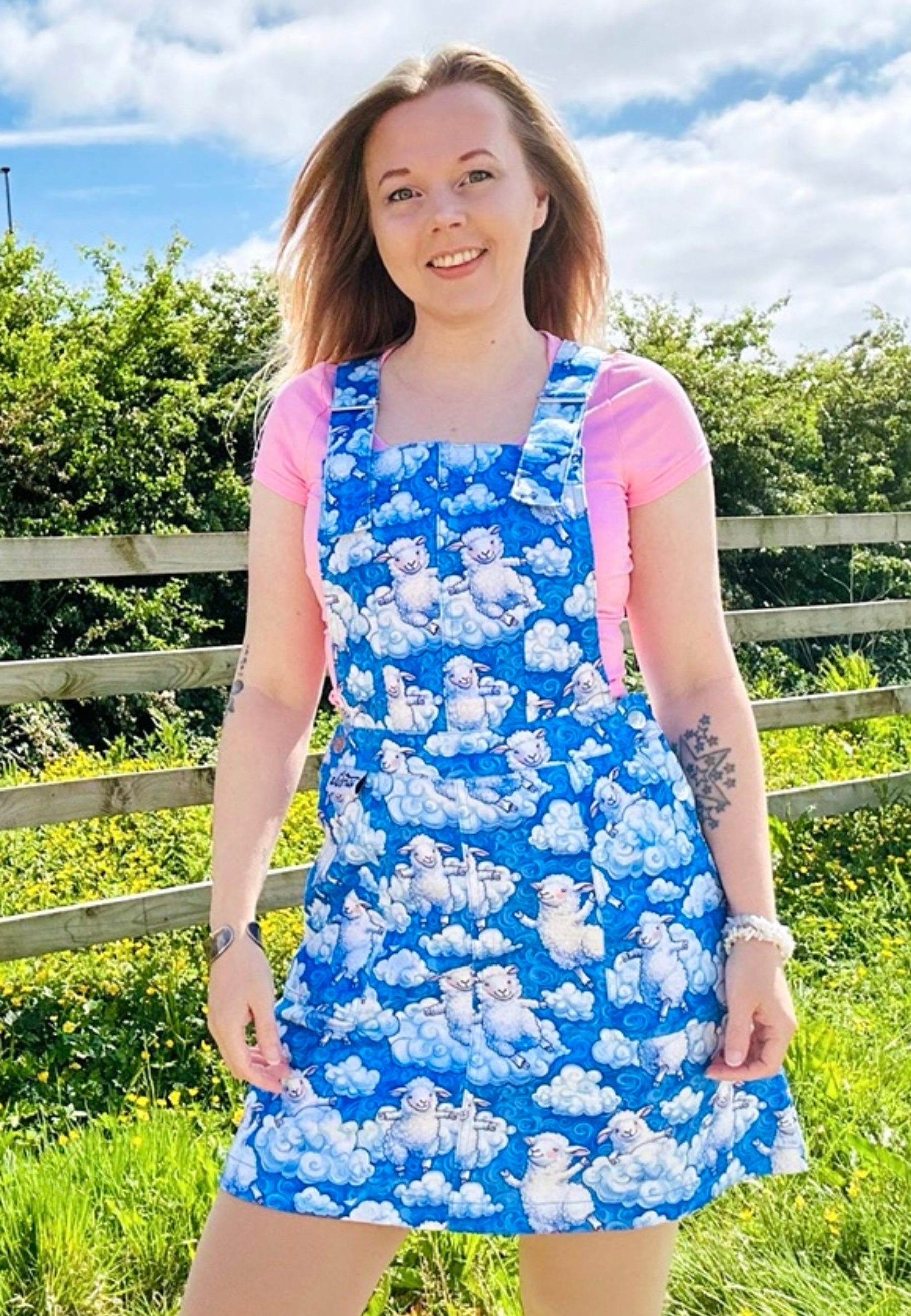 Woman wearing a blue pinafore dress with a beautiful cloud and sheep pattern. Woman is outdoors under blue cloud sky. 