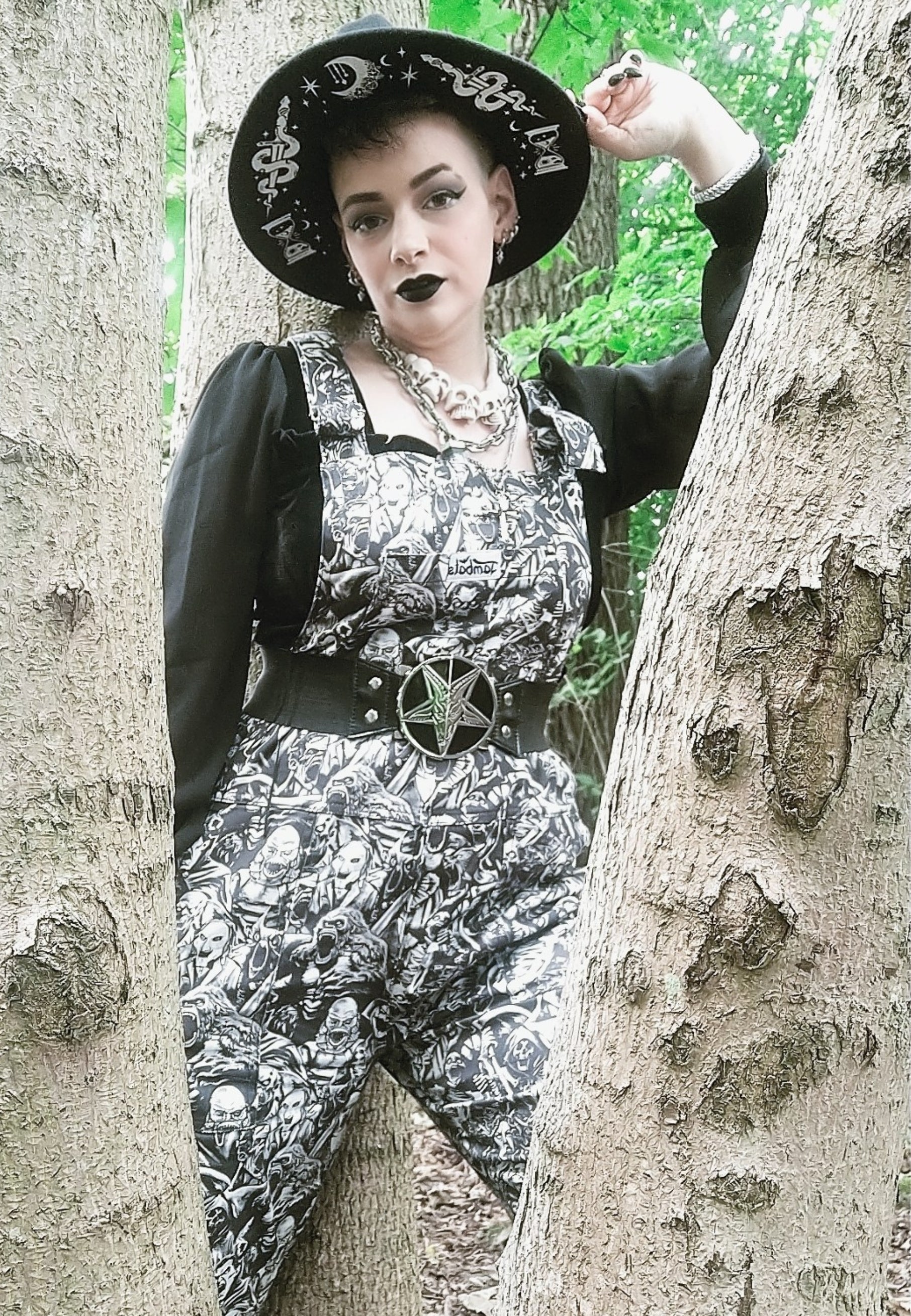 An alternative styled woman  wearing black and white grayscale print dungarees over a black mesh top. Dungaree print is old school and modern monsters, horror characters and cryptids in an old horror comic style illustration. They are all screaming. Woman is wearing a gothic hat and pentagram star belt.