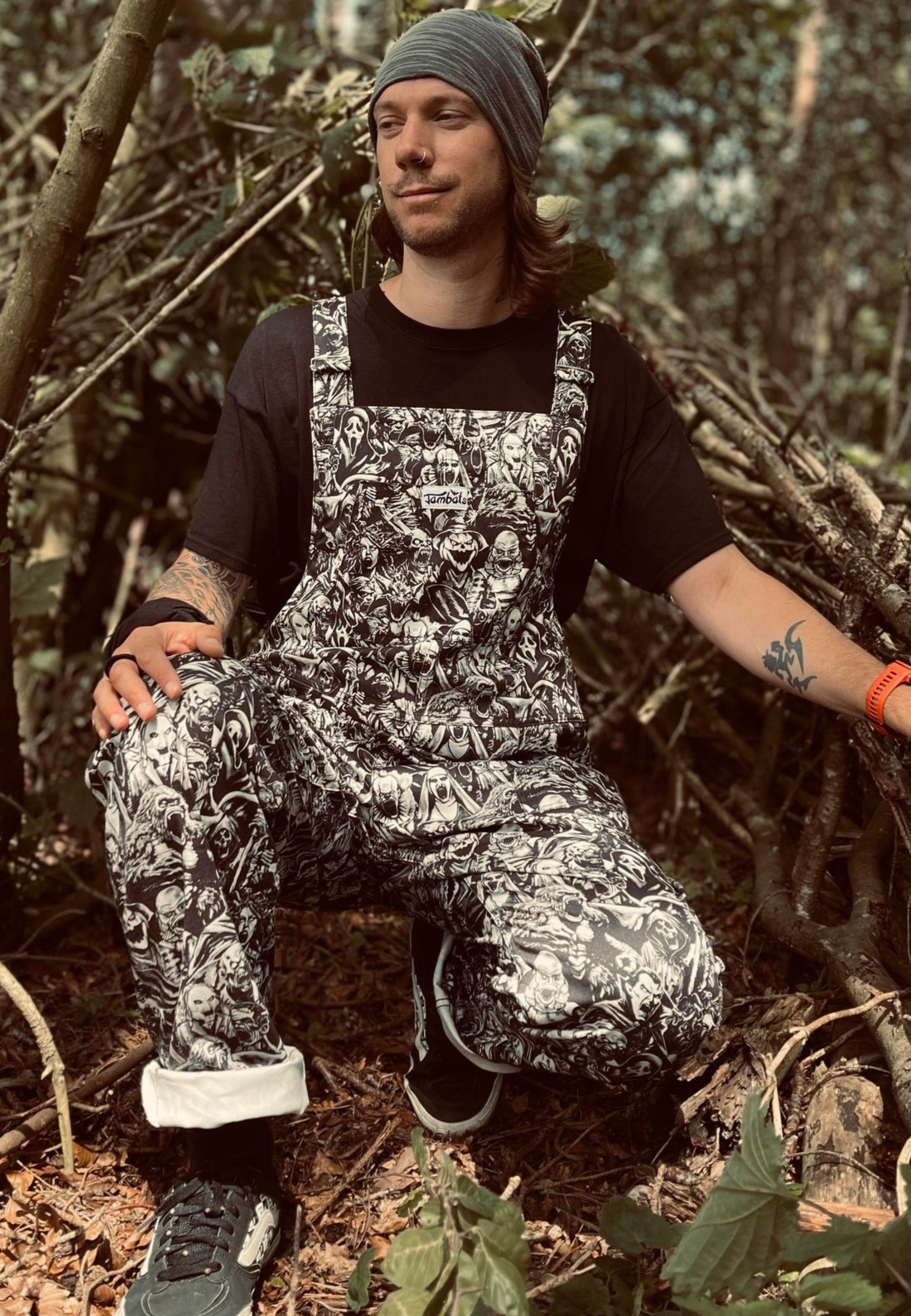 An alternative styled man  outdoors wearing black and white grayscale print dungarees over a black mesh top. Dungaree print is old school and modern monsters, horror characters and cryptids in an old horror comic style illustration. They are all screaming.