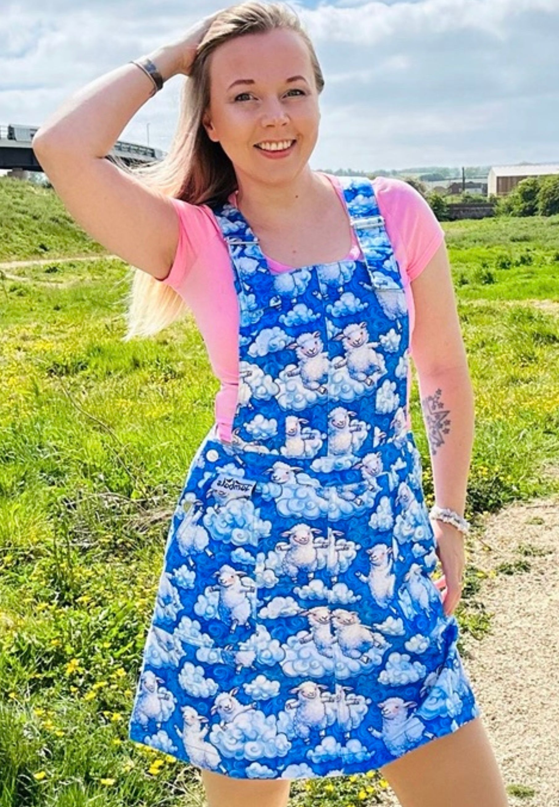 Woman wearing a blue pinafore dress with a beautiful cloud and sheep pattern. Woman is outdoors under blue soft fluffy cloud sky. Also wearing pink trainers. 