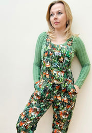 A woman wearing green Cat Nap dungarees with a pattern of various sleeping cats among houseplants. The dungarees have chest, hip, and bum pockets and feature a slider buckle fastener.
