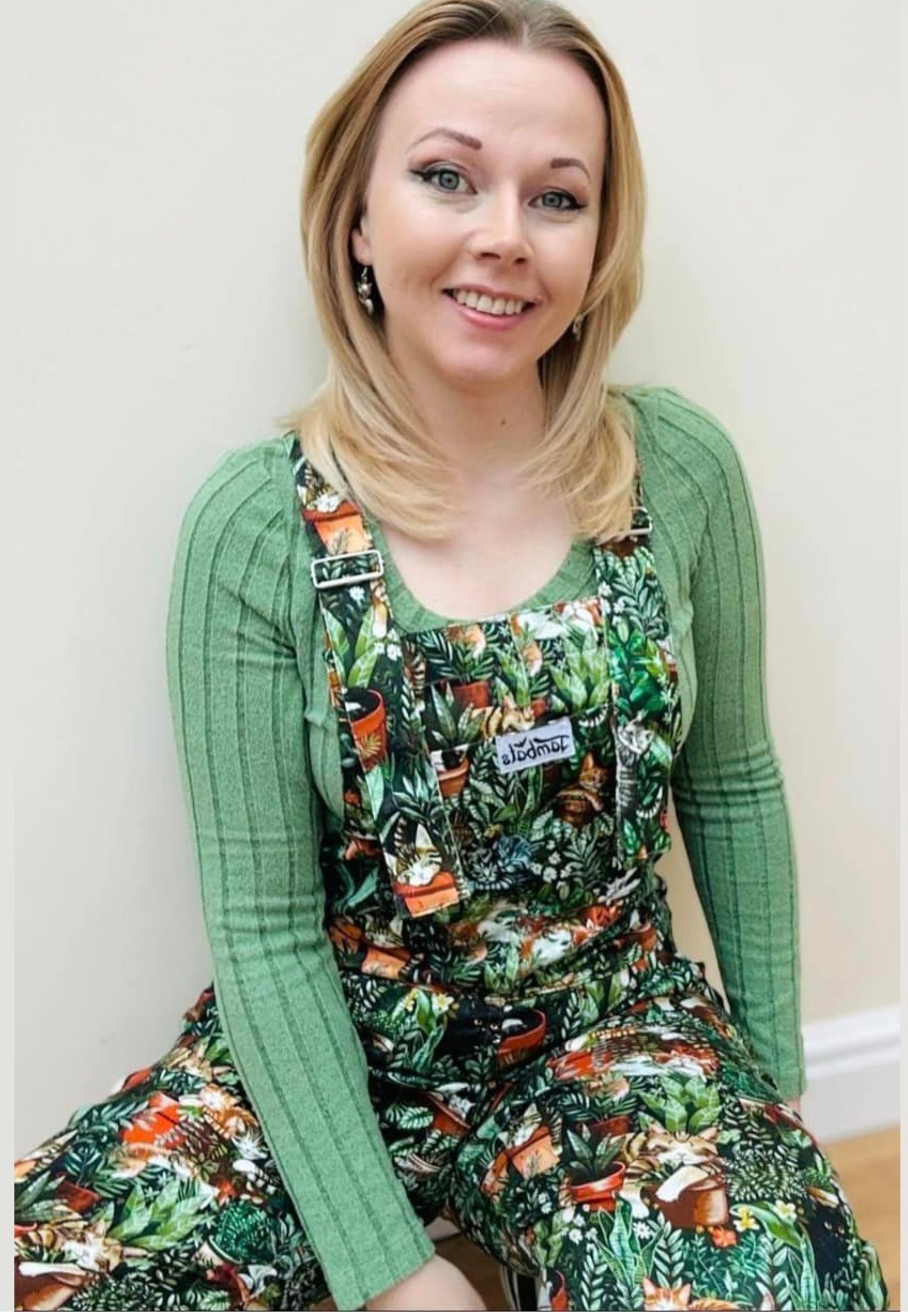A woman wearing green Cat Nap dungarees with a pattern of various sleeping cats among houseplants. The dungarees have chest, hip, and bum pockets and feature a slider buckle fastener.