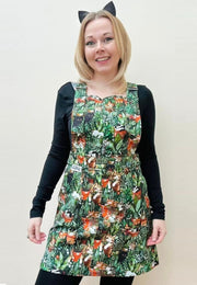 A woman wearing a multi coloured pinafore dress with a pattern of cats and houseplants, paired with a black long-sleeved top and woman is wearing cute cat ears hairband. 