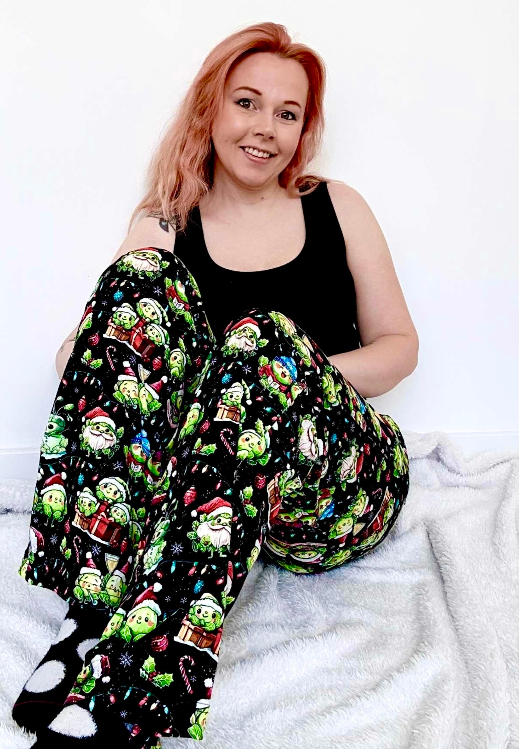 Woman wearing a black tank top and colorful patterned lounge pants sitting on a white surface. Pants have the cutest Christmas Sprouts print