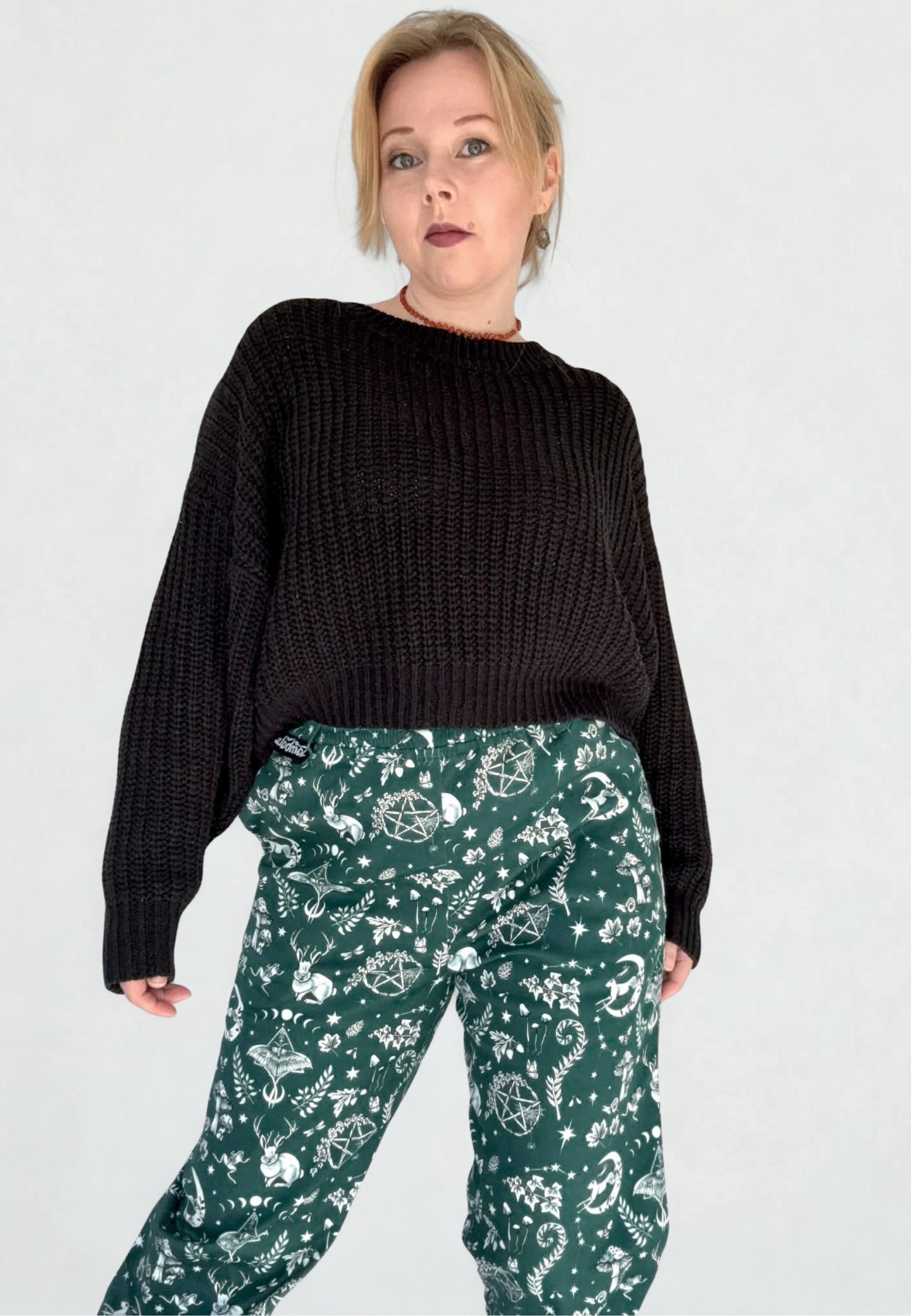Woman in glasses wearing a black slouchy jumper and green trousers with a Magick print on, standing against a white background