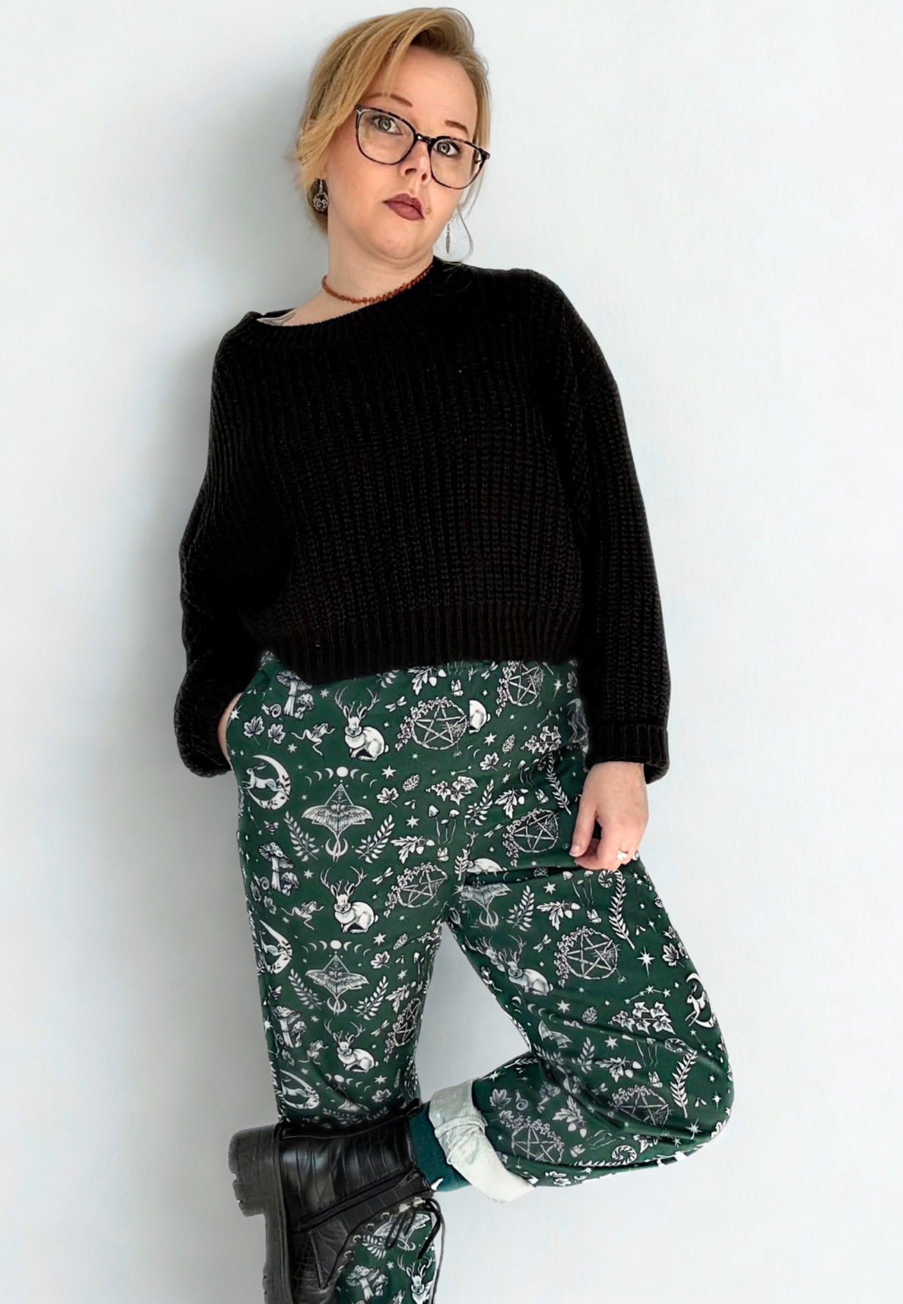 Woman in glasses wearing a black slouchy jumper and green trousers with a Magick print on, standing against a white background