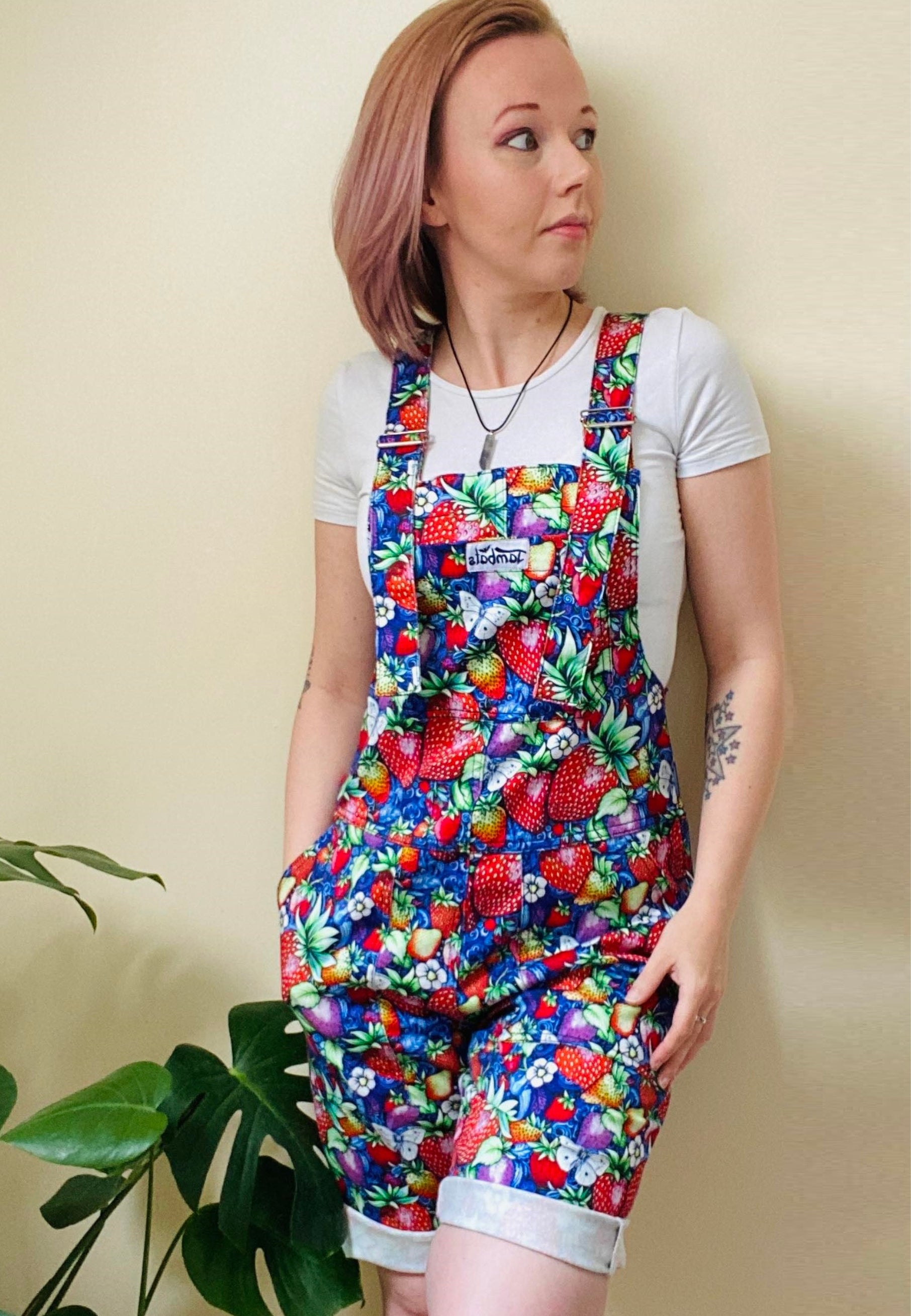 A person wearing a vibrant multicolored floral dungaree shortie with a blue paisley pattern. They have their hands in the pockets and are standing against a neutral background with a plant to the side.