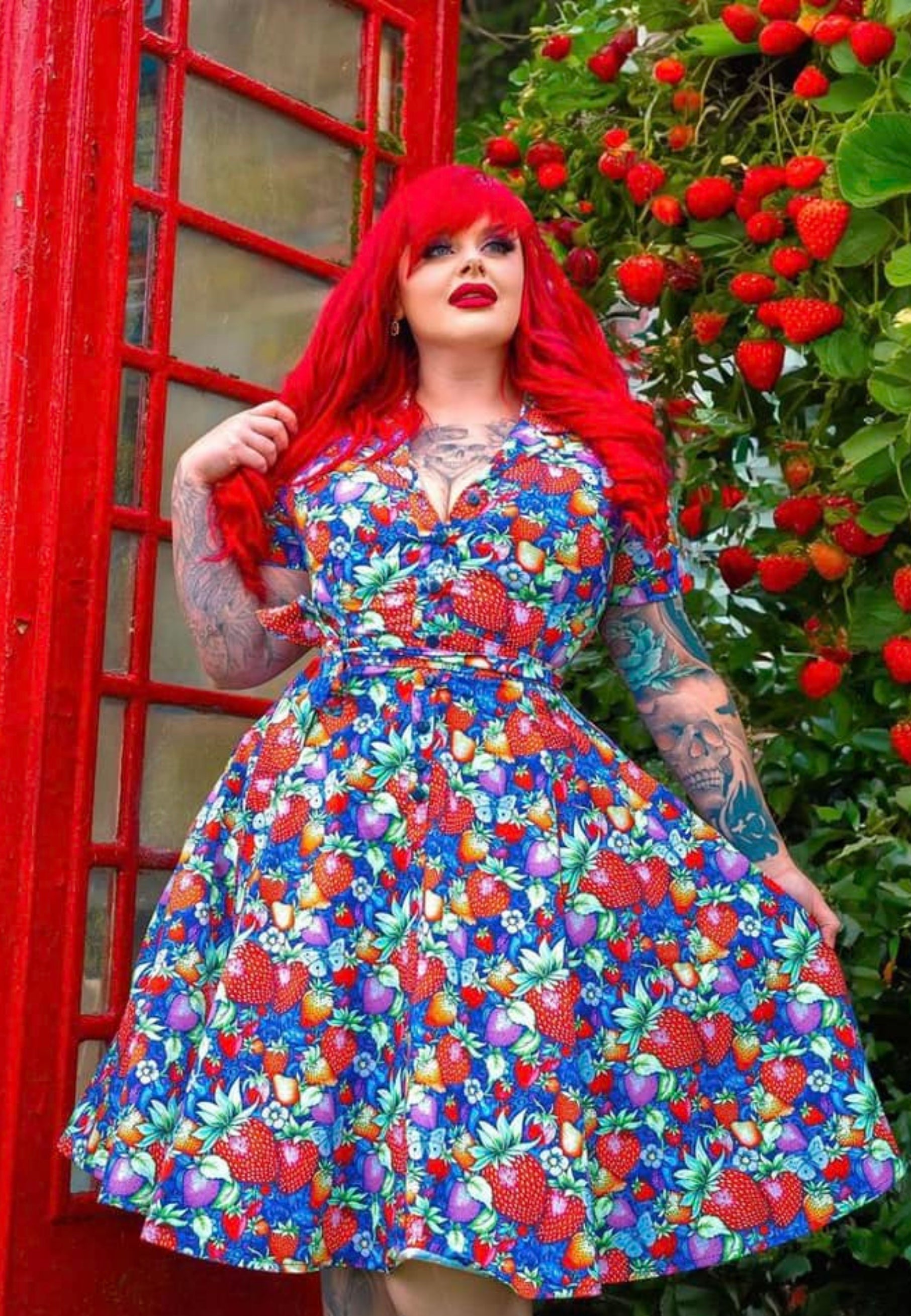 A woman with long red hair wearing a vibrant retro shirt dress with a strawberry and paisley pattern, standing in front of an iconic red British telephone box with strawberry plants.