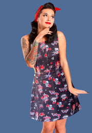 A woman modeling a black stretch cotton twill pinafore dress with a traditional tattoo print, featuring deep hip pockets and side button fastenings.