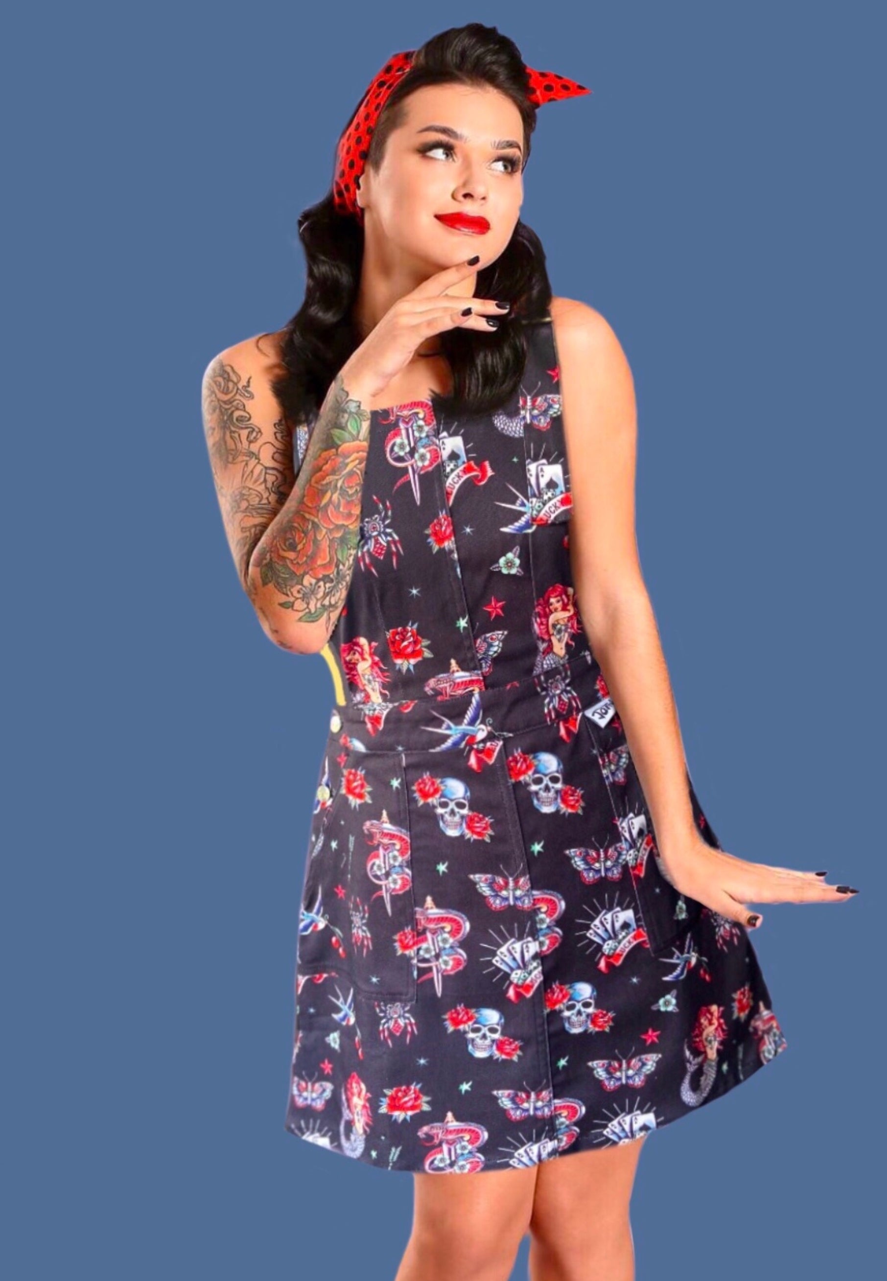 A woman modeling a black stretch cotton twill pinafore dress with a traditional tattoo print, featuring deep hip pockets and side button fastenings.