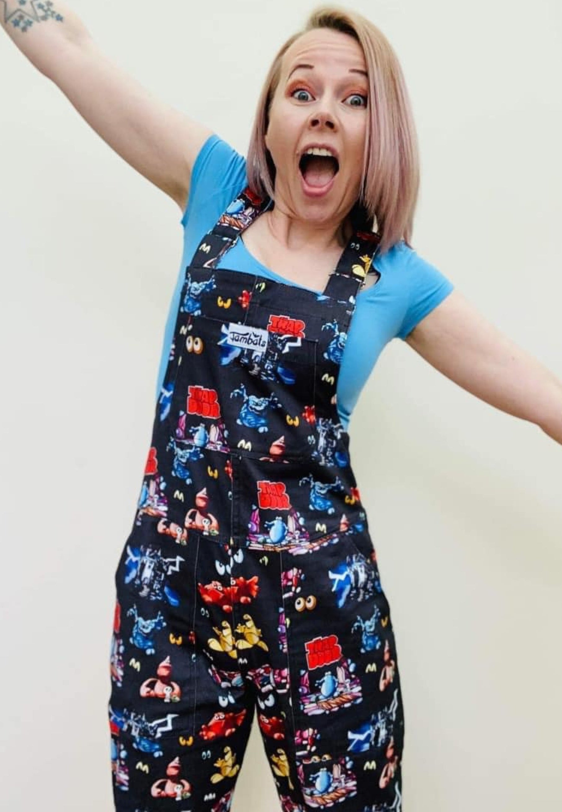A woman wearing a pair of Trap Door dungarees with a print of characters frpm the 80's 90's childrens tv show. The dungarees are predominantly dark-colored with colorful character illustrations.