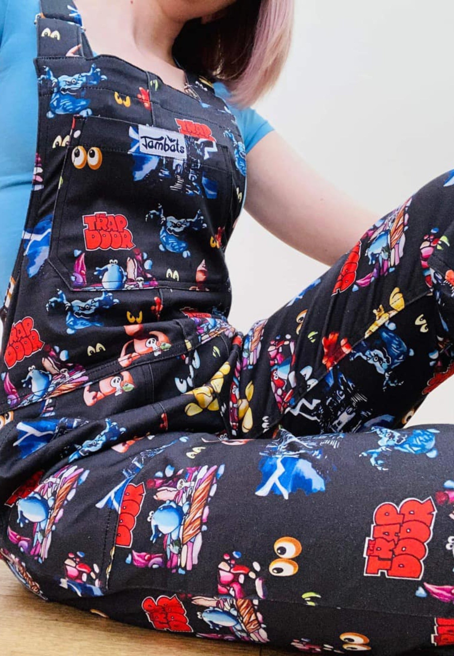 A woman wearing a pair of Trap Door dungarees with a print of characters frpm the 80's 90's childrens tv show. The dungarees are predominantly dark-colored with colorful character illustrations. Close up image of the dungarees.