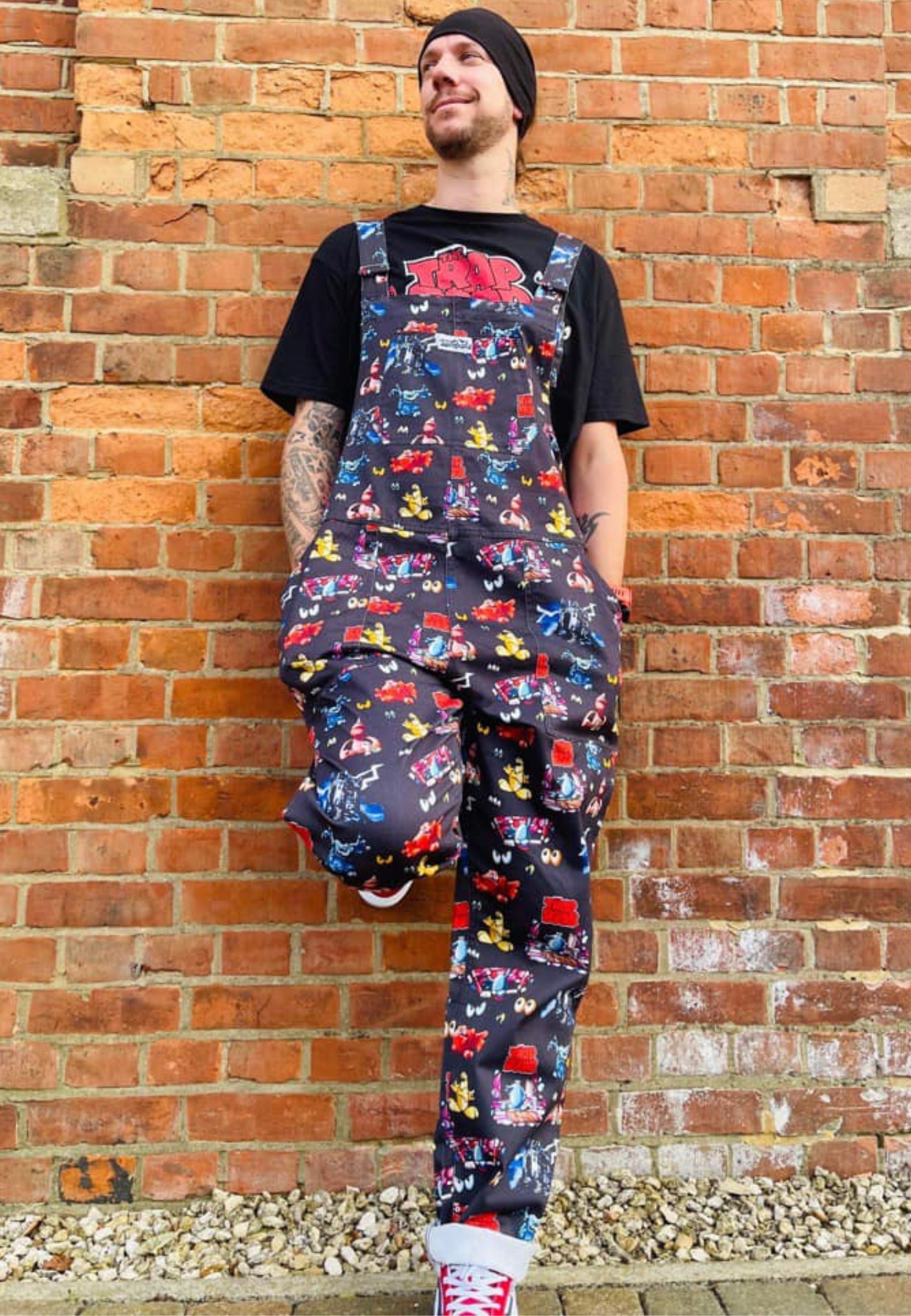 A man stood next to a wall wearing a pair of Trap Door dungarees with a print of characters frpm the 80's 90's childrens tv show. The dungarees are predominantly dark-colored with colorful character illustrations.
