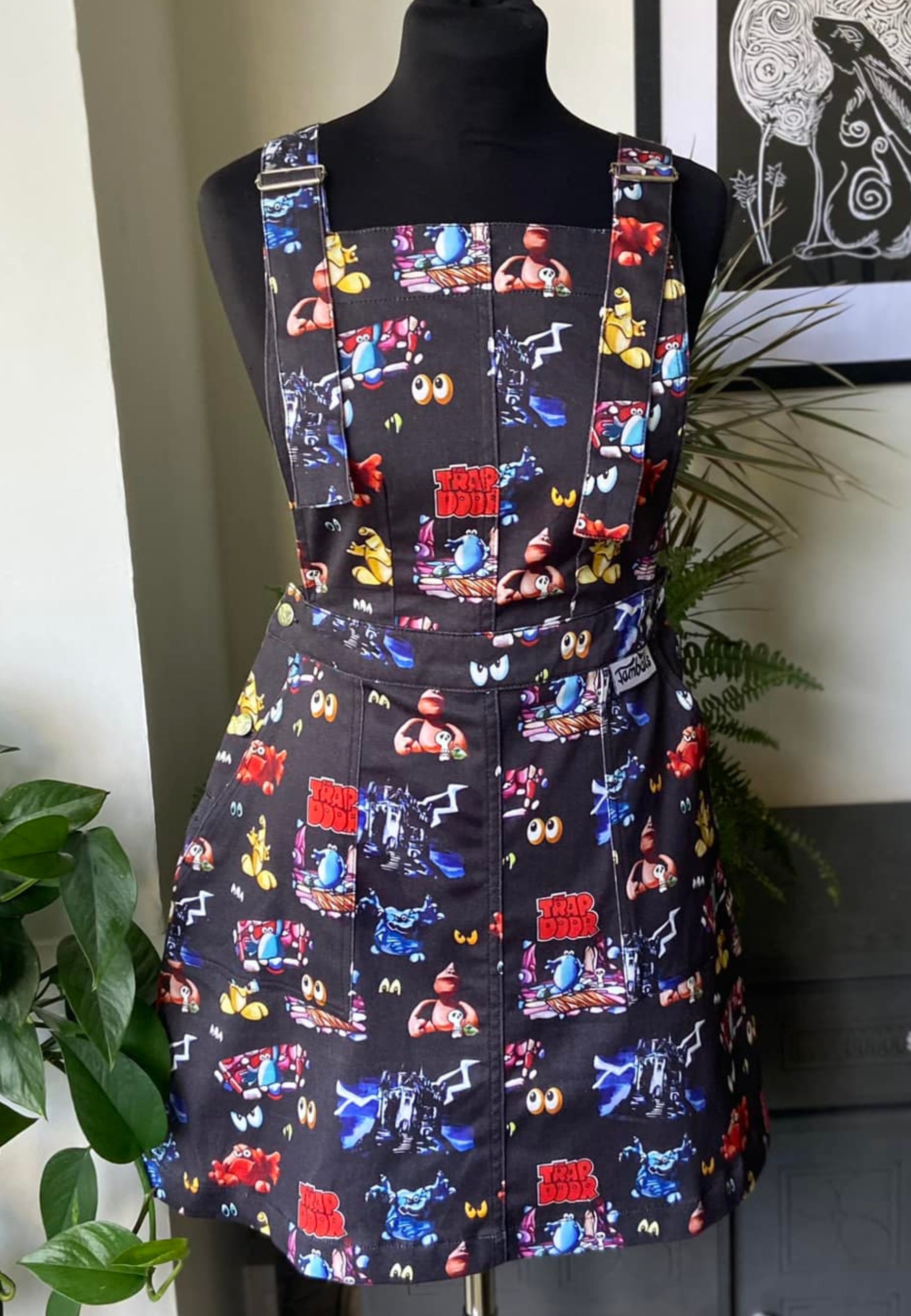A black pinafore dress displayed on a mannequin featuring a colorful pattern with various characters and text from the cult 80's tv progremme Trap Door.