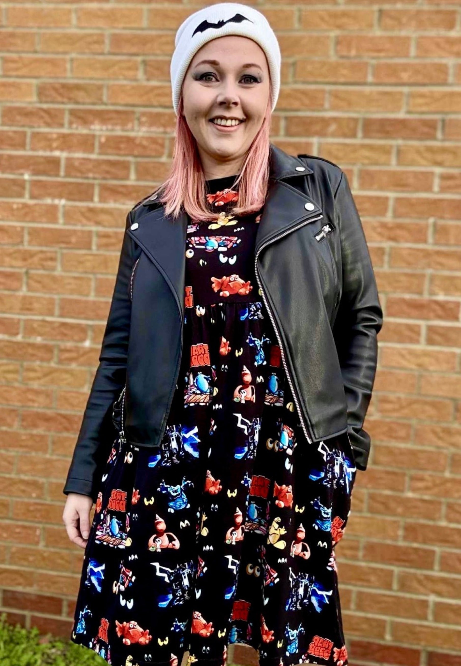 Woman wearing a black Trap Door print dress, black leather jacket, and white beanie in front of a brick wall.