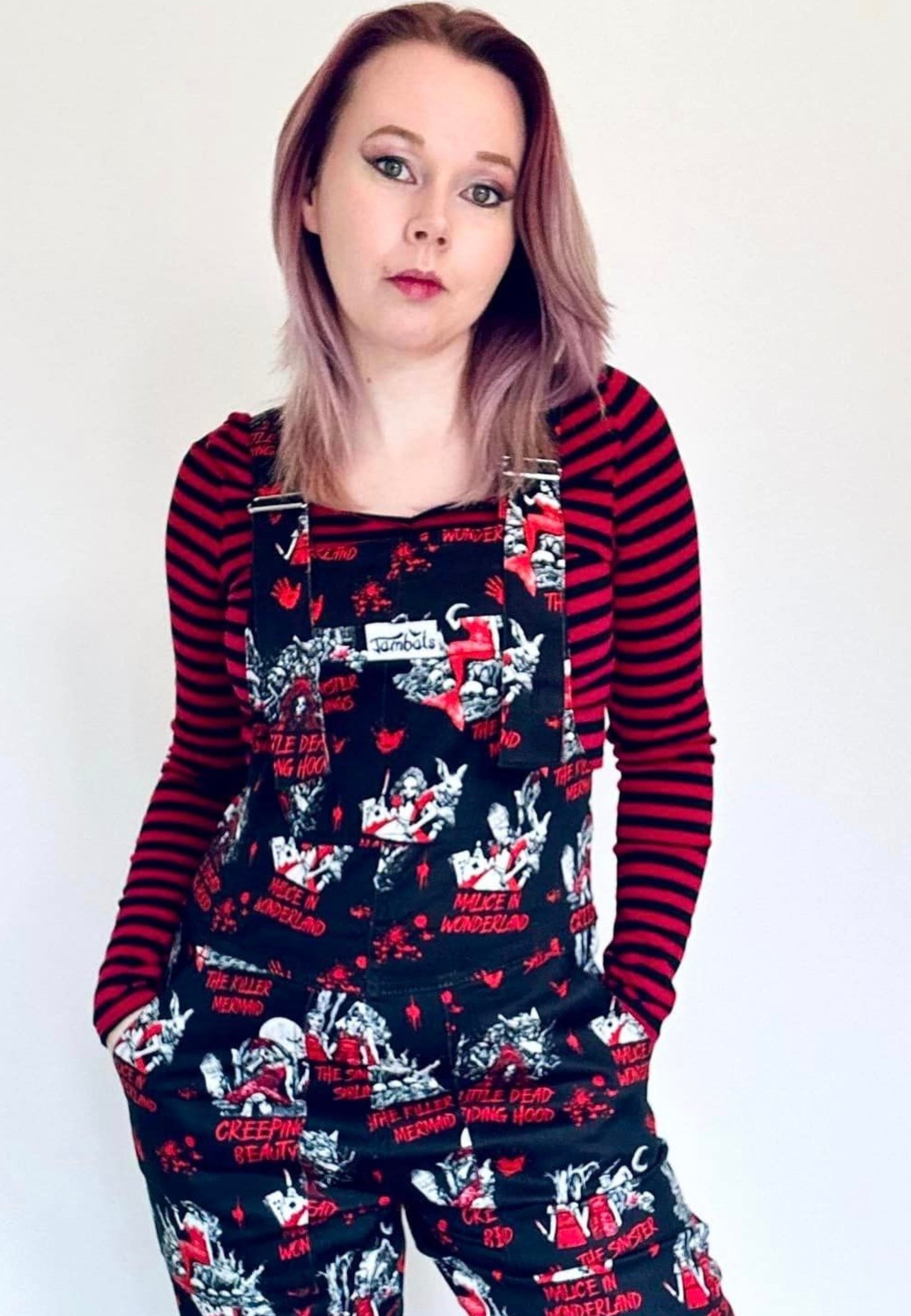 A woman wearing a pair of Twisted Tales print dungarees, featuring character illustrations of dark fairytales in a dark whimsical pattern on a black background. Also wearing a black and red top. 