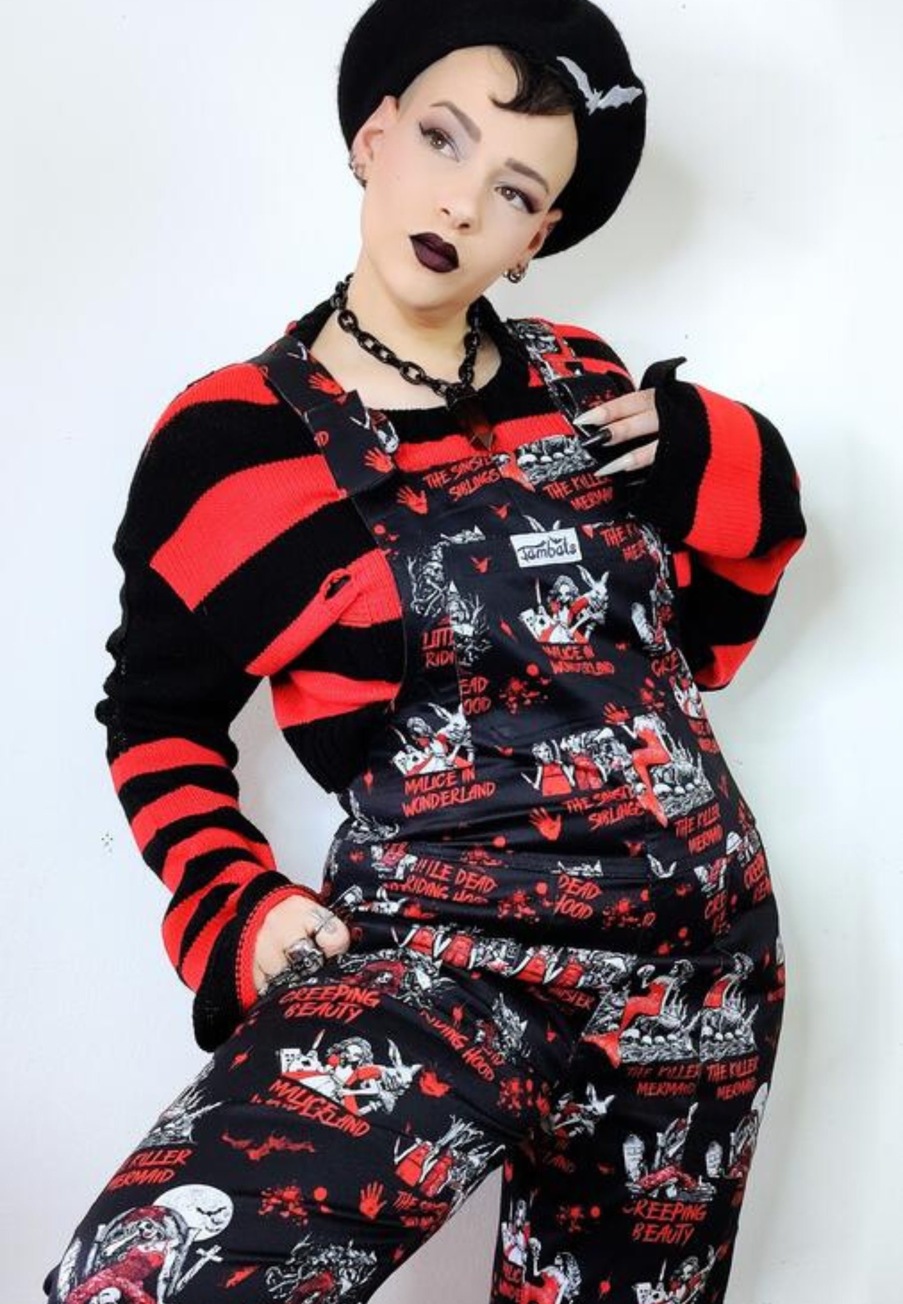 A woman wearing a pair of Twisted Tales print dungarees, featuring character illustrations of dark fairytales in a dark whimsical pattern on a black background. Also wearing a black and red striped to and a beret. Alternative styling. 