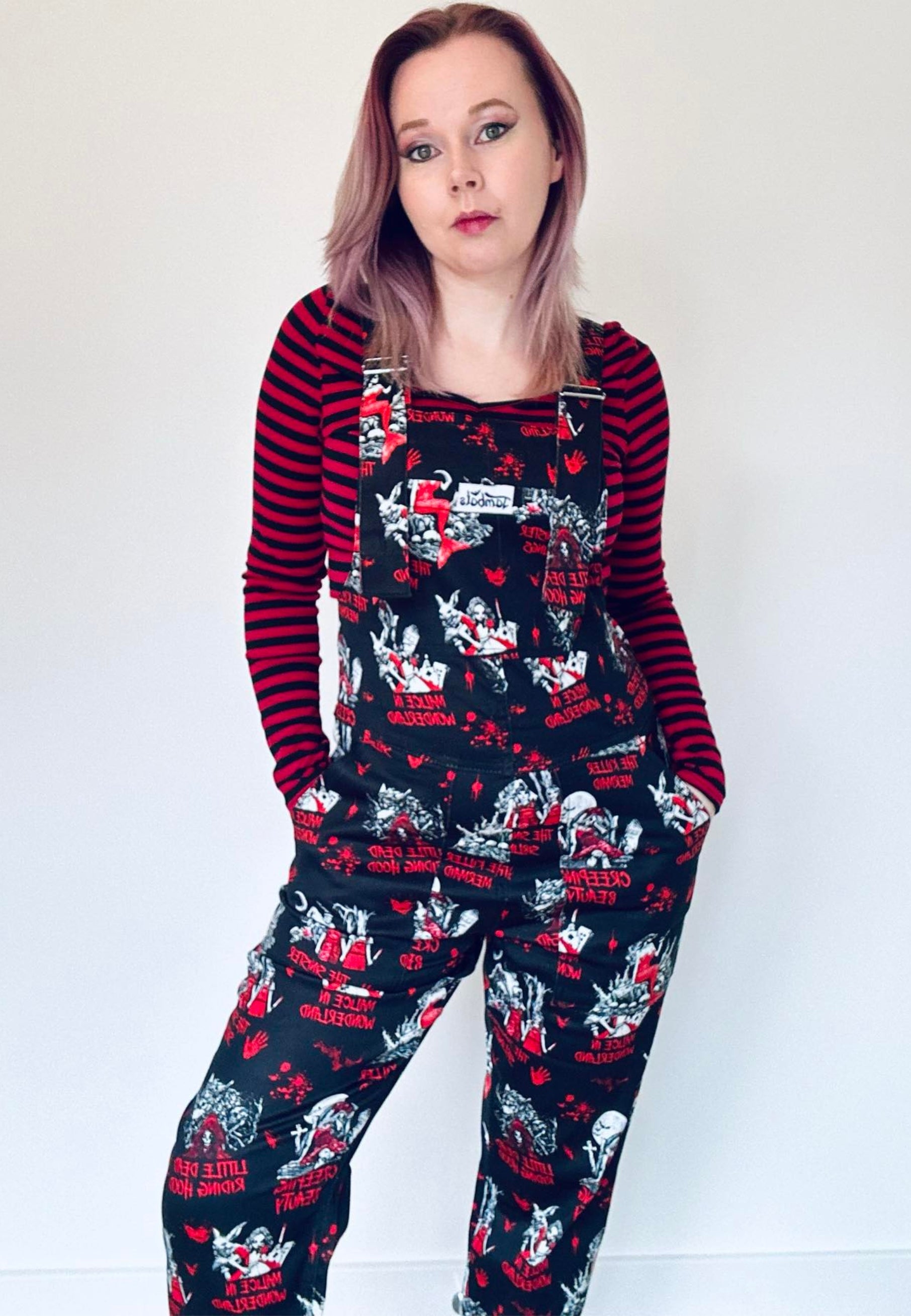 A person wearing a pair of Twisted Tales print dungarees, featuring character illustrations of dark fairytales in a dark whimsical pattern on a black background. 