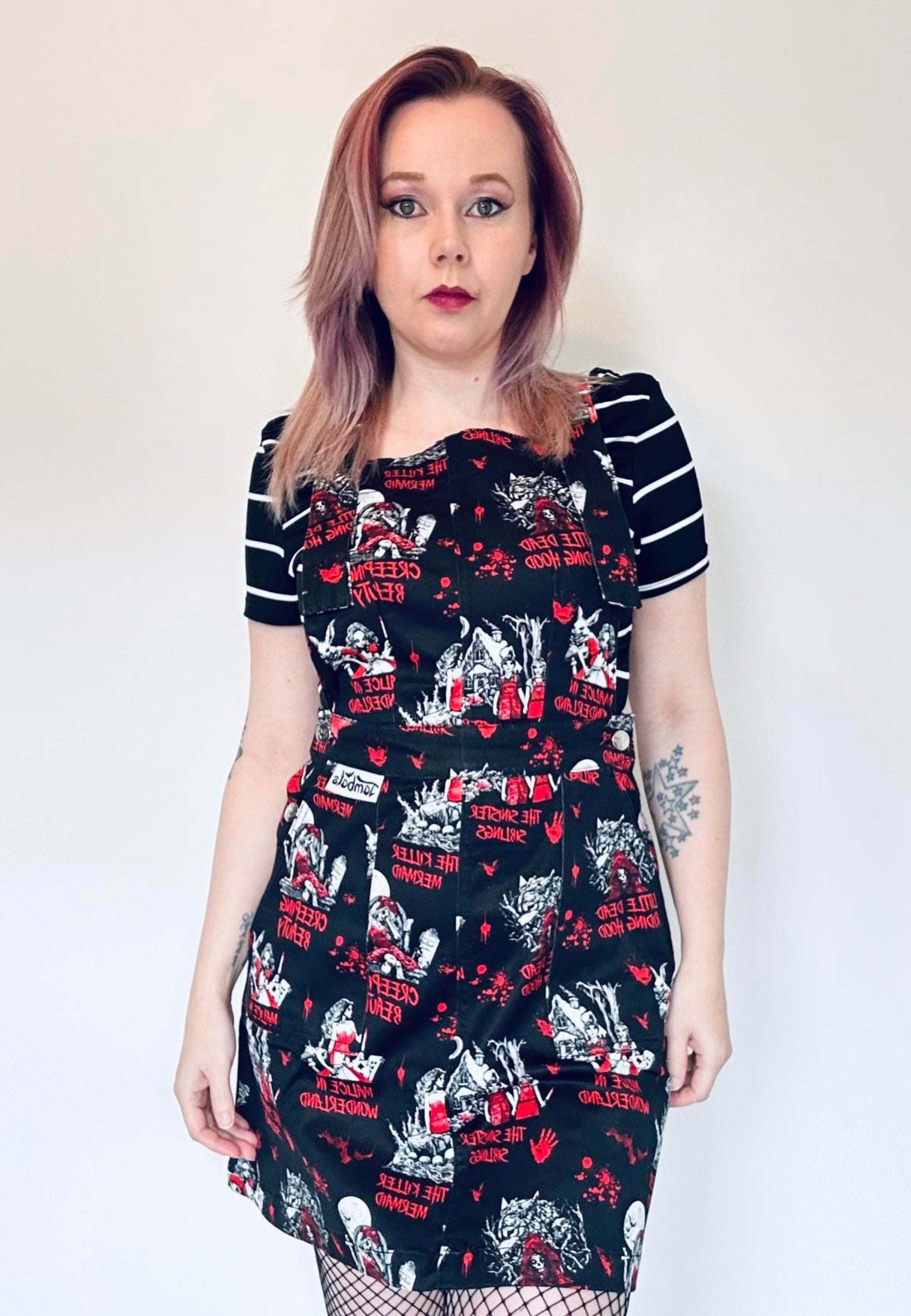 A woman wearing a black pinafore dress with a red and white character-based print, paired with a striped top underneath. Print on the pinafore is Jambats Twisted Tales which is a goth, horror print of dark fairytales. 