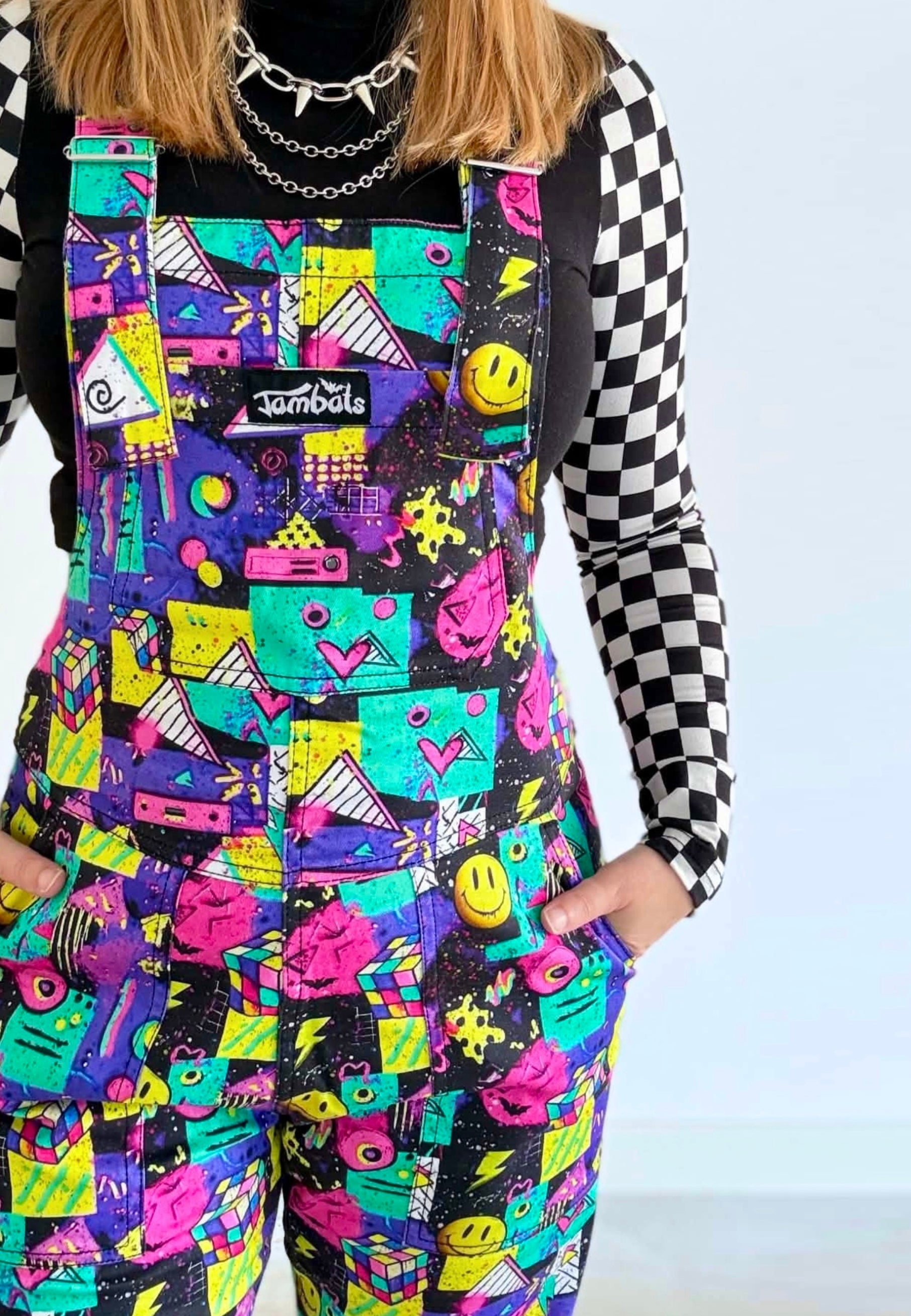 A woman wearing bright multicolour 80s 90s print dungarees overalls with a black-and-white checkered long-sleeve shirt underneath. The overalls feature a colourful retro graffiti style print with smiley faces, hearts, stars, and geometric shapes on a black base.