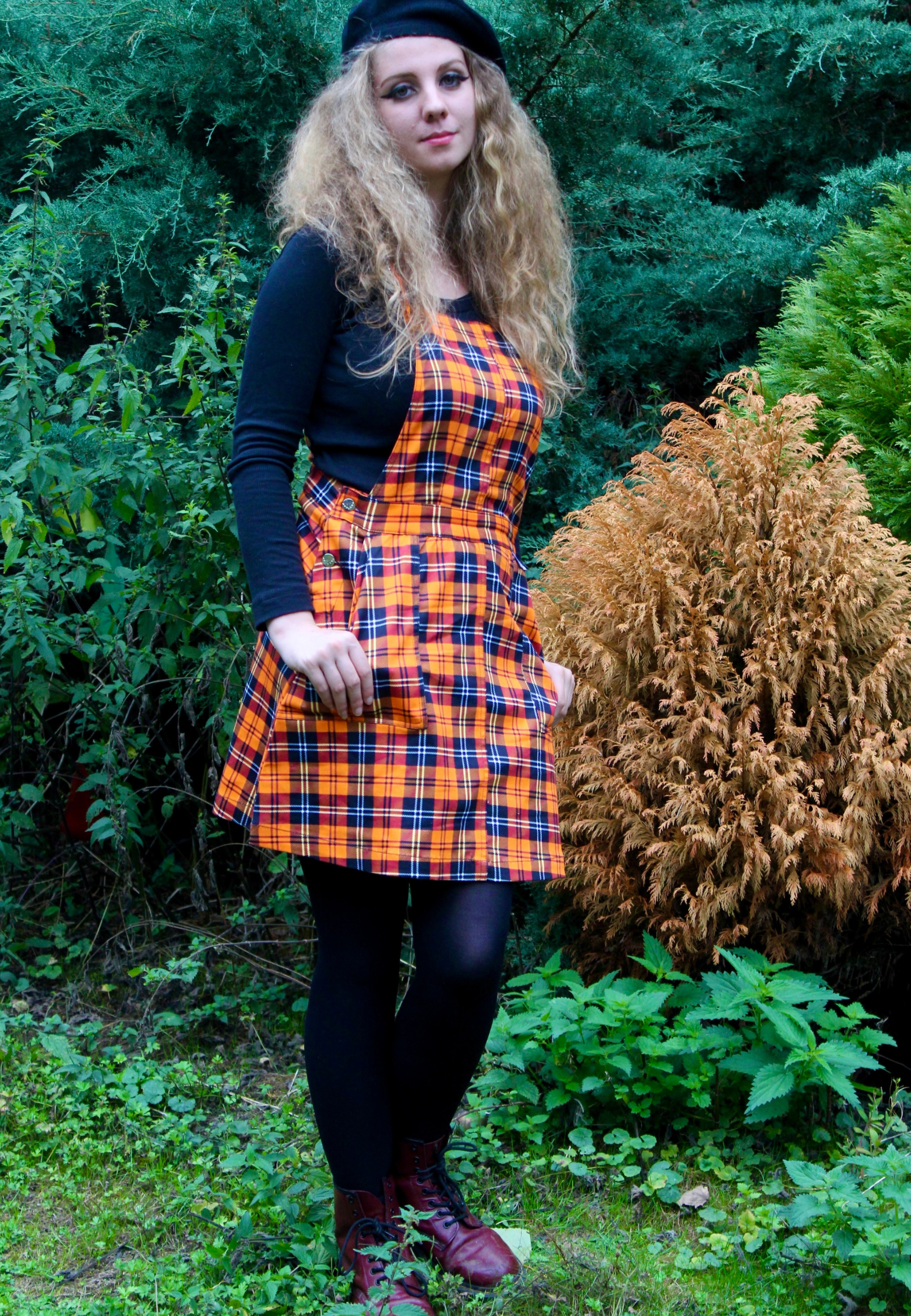 Woman wearing an orange tartan pinafore dress over a black sweater in a forest wearing a black beret.