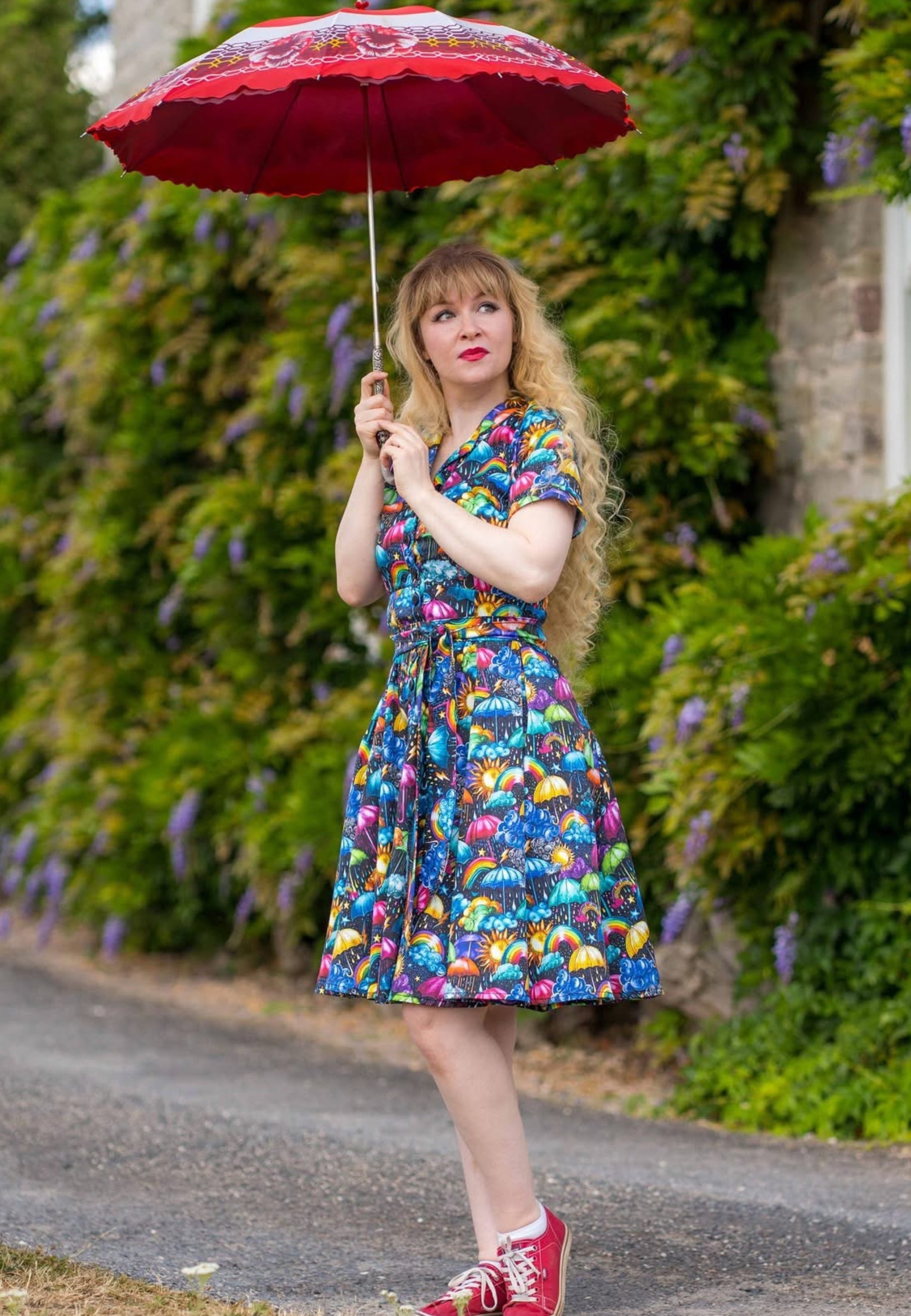 A woman stands outdoors holding an umbrella, wearing a beautiful, vibrant, whimsical weather patterned dress with red pumps. The background shows a garden wall with green foliage and purple flower