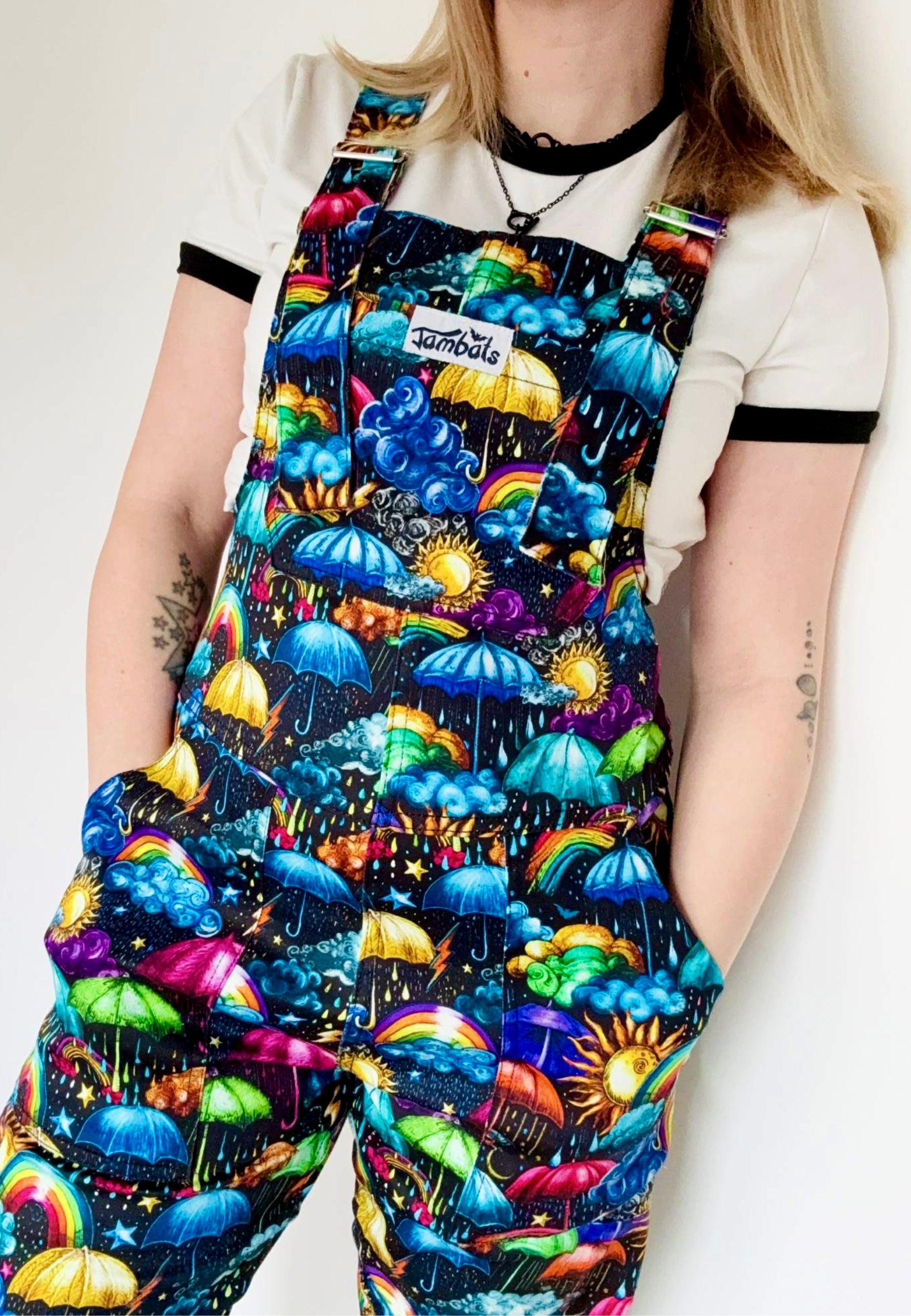 Close up studio photo of a woman wearing dungarees with a vibrant weather print. Whimsical fabric print of umbrellas, clouds, rain, sunshine, rainbows, storm clouds. 