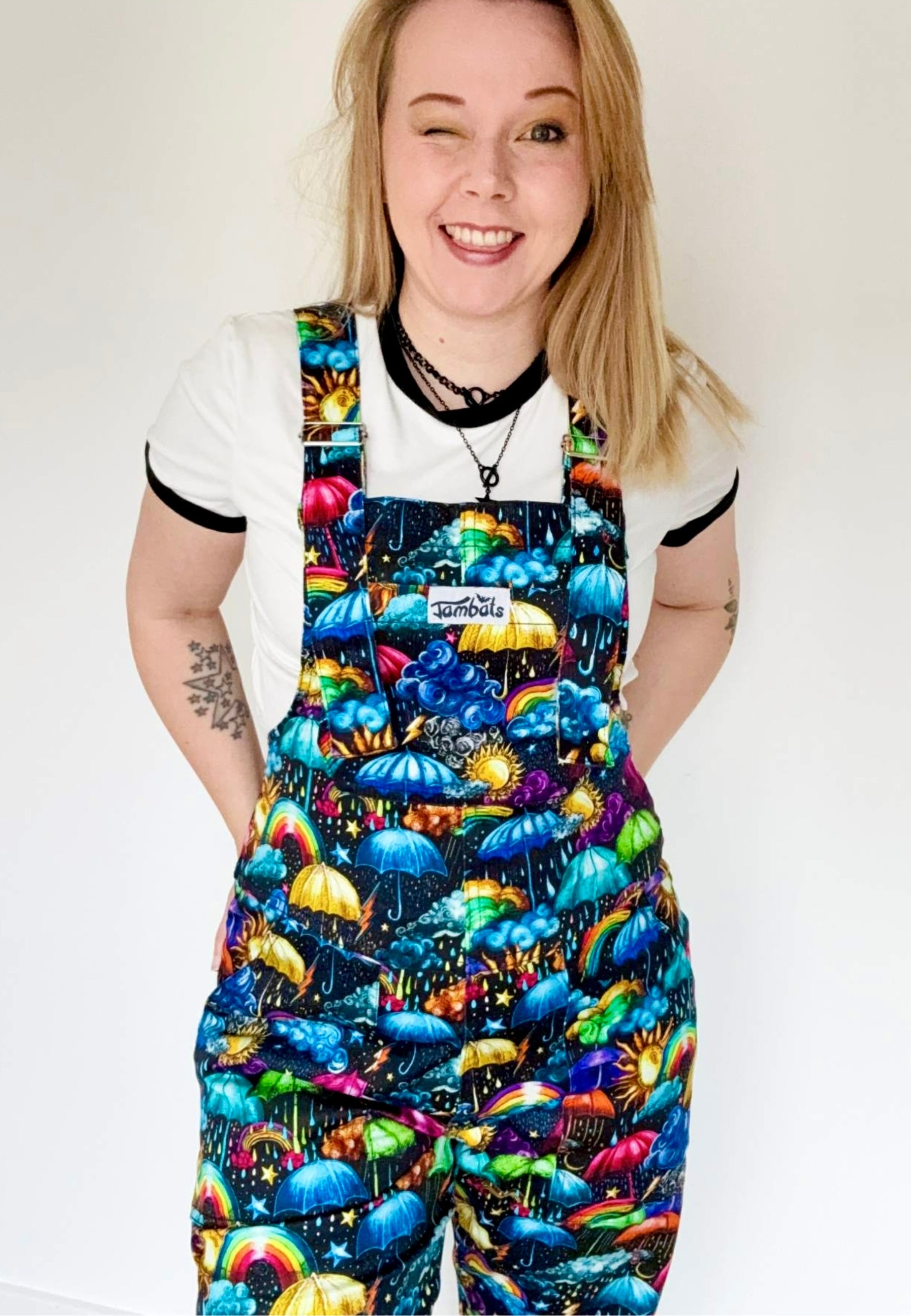 Woman smiling to camera and wearing dungarees with a vibrant weather print. Whimsical fabric print of umbrellas, clouds, rain, sunshine, rainbows, storm clouds. Studio photo. 
