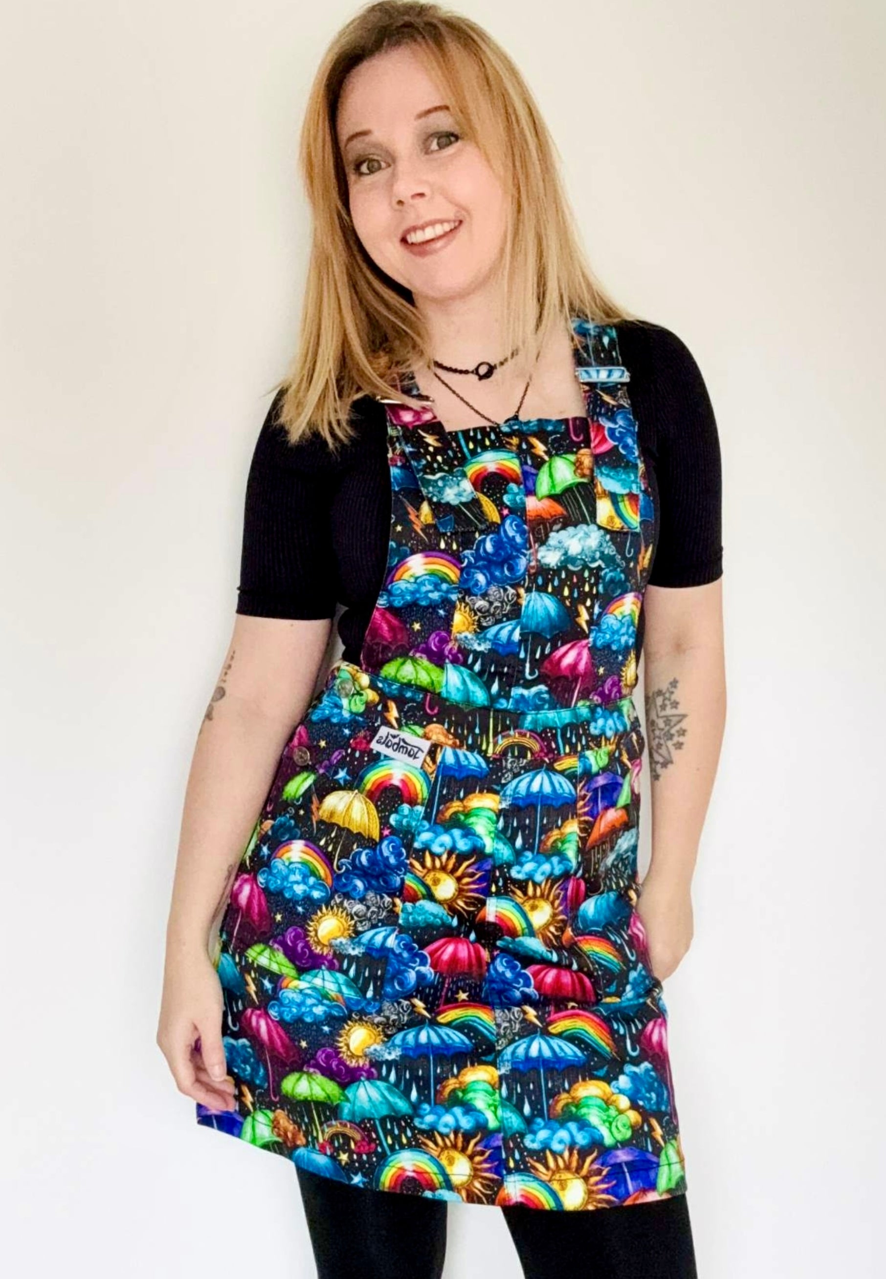 Woman wearing a pinafore dress with a vibrant weather print. Whimsical fabric print of umbrellas, clouds, rain, sunshine, rainbows, storm clouds. Studio photo. 
