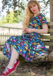 A woman sits on a bench outdoors, wearing a beautiful, vibrant, whimsical weather patterned dress with red pumps. The background shows a garden with green foliage. 