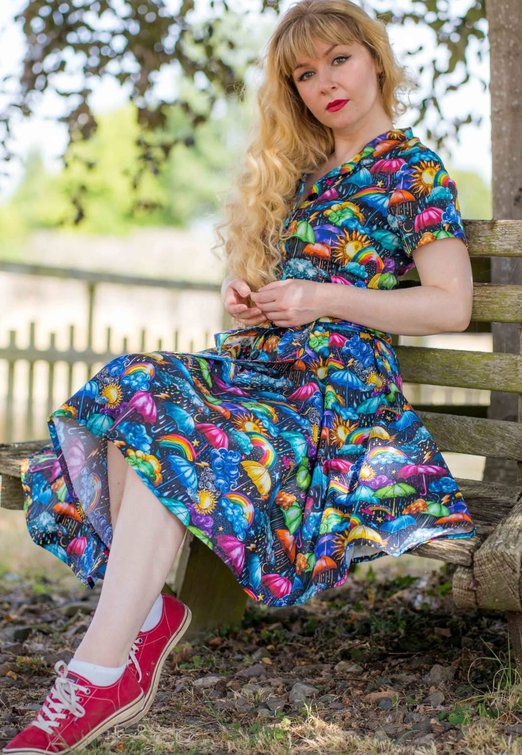 A woman sits on a bench outdoors, wearing a beautiful, vibrant, whimsical weather patterned dress with red pumps. The background shows a garden with green foliage. 