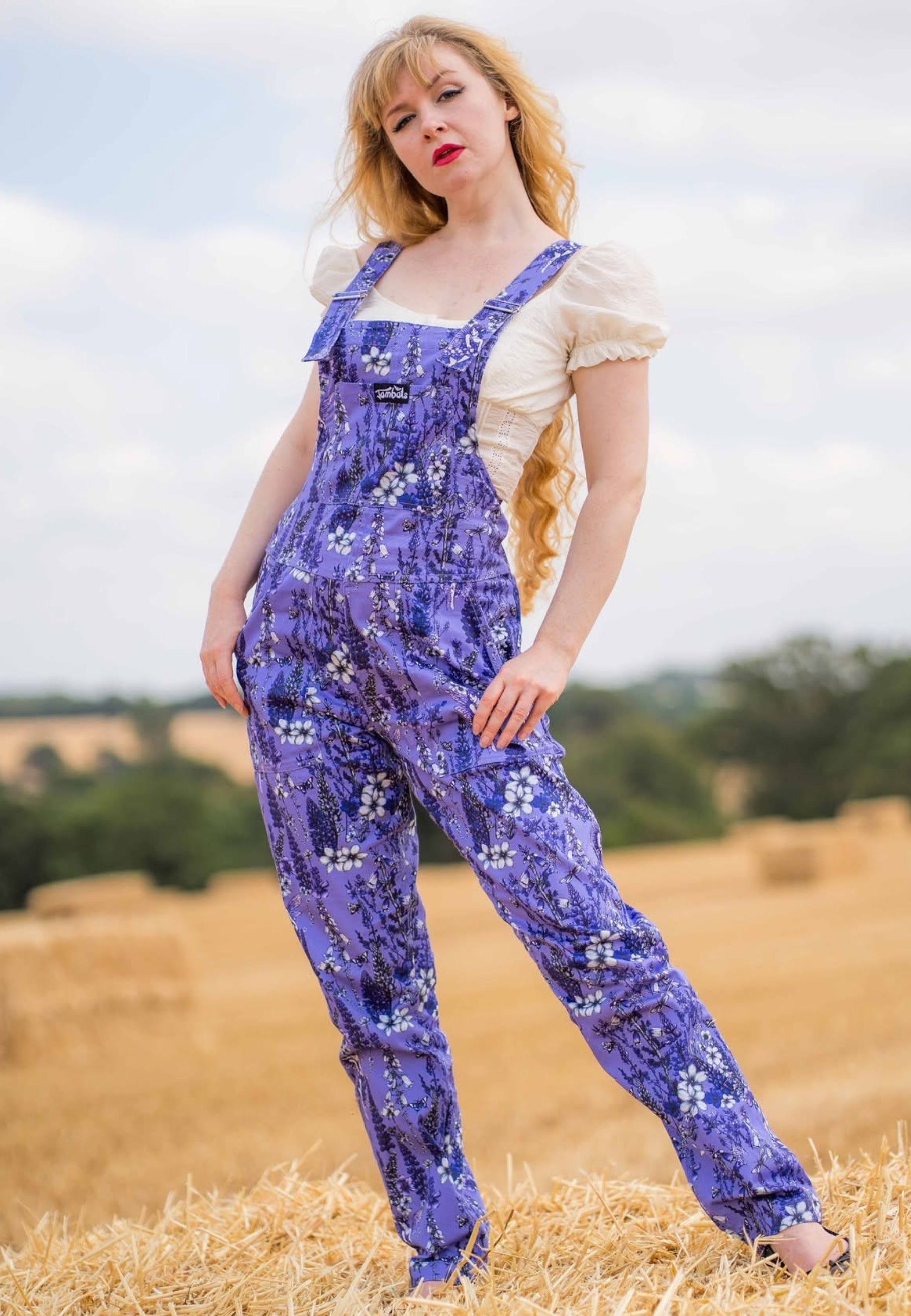 A woman standing in a field of corn, wearing purple floral print overalls dungarees over a cream puff-sleeve top.