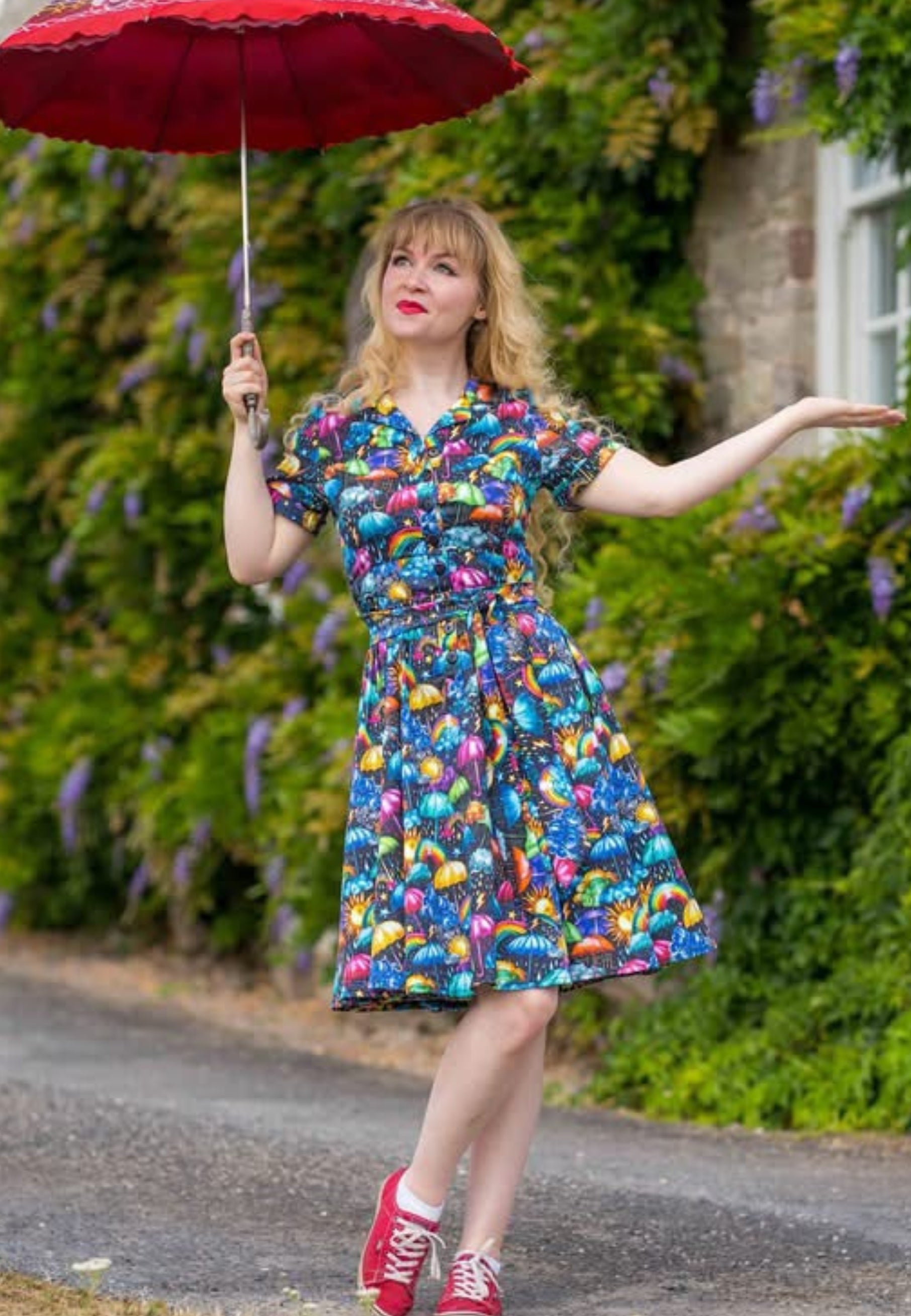 A woman stands outdoors holding an umbrella, wearing a beautiful, vibrant, whimsical weather patterned dress with red pumps. The background shows a garden wall with green foliage and purple flowers.
