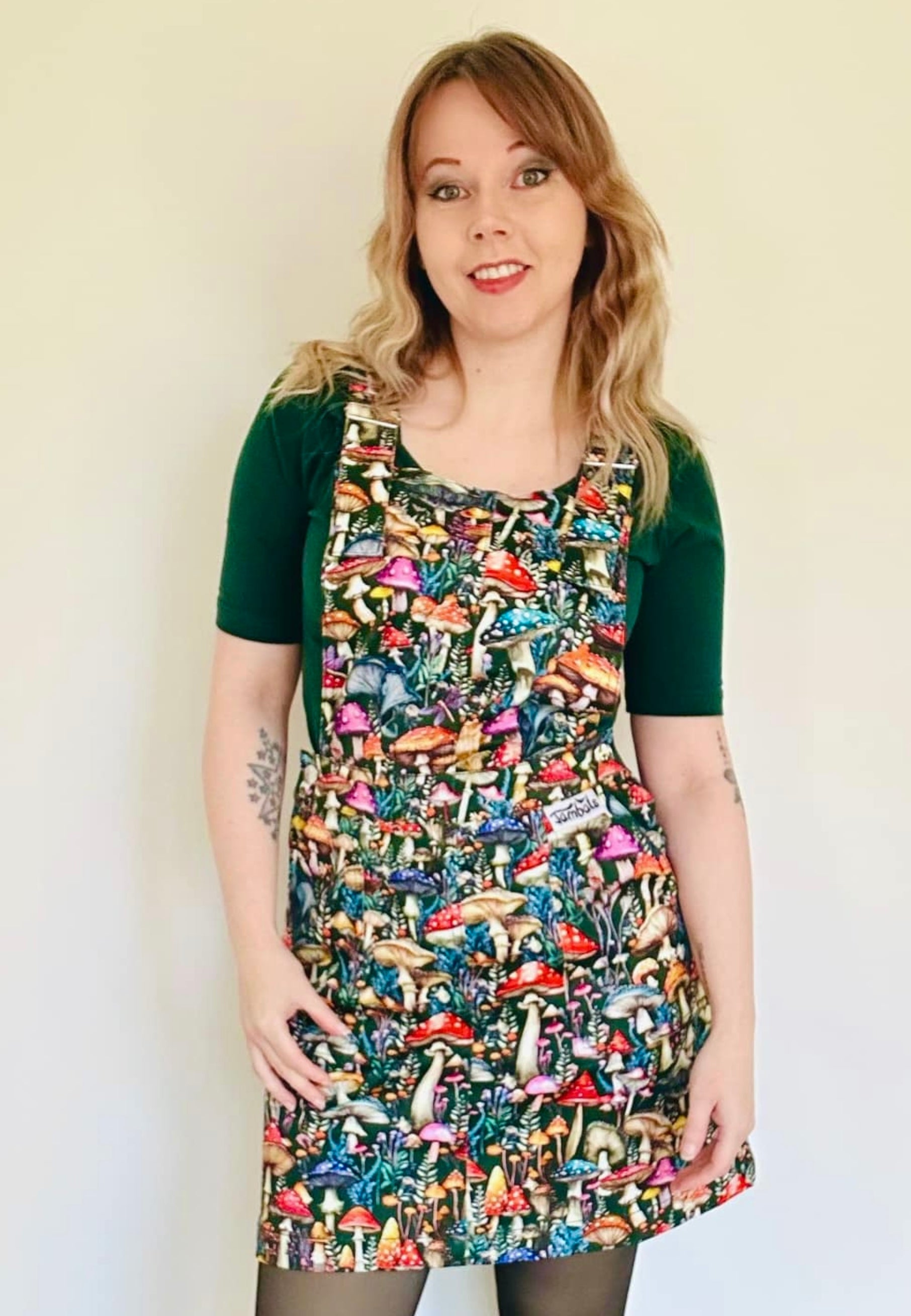 A woman wearing a dark green pinafore dress with a vibrant mushroom print with mushrooms and toadstools. Studio photo on a plain white background. 