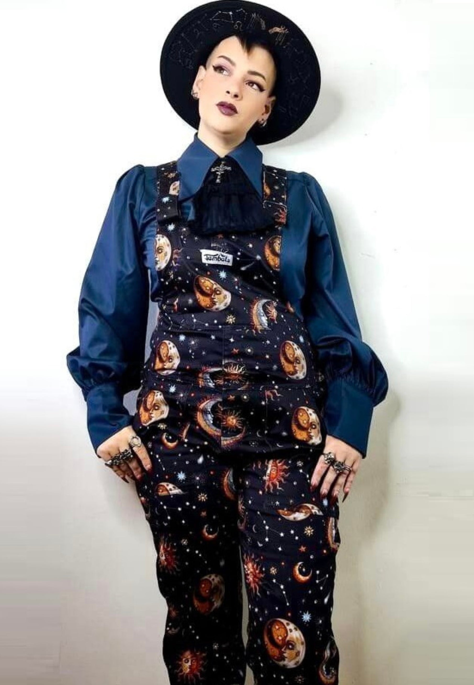 A woman wearing a regular fit, celestial print dungarees with a chest pocket, hip pockets, and a bum pocket. The dungarees are black with a unique fabric print featuring moons and stars.