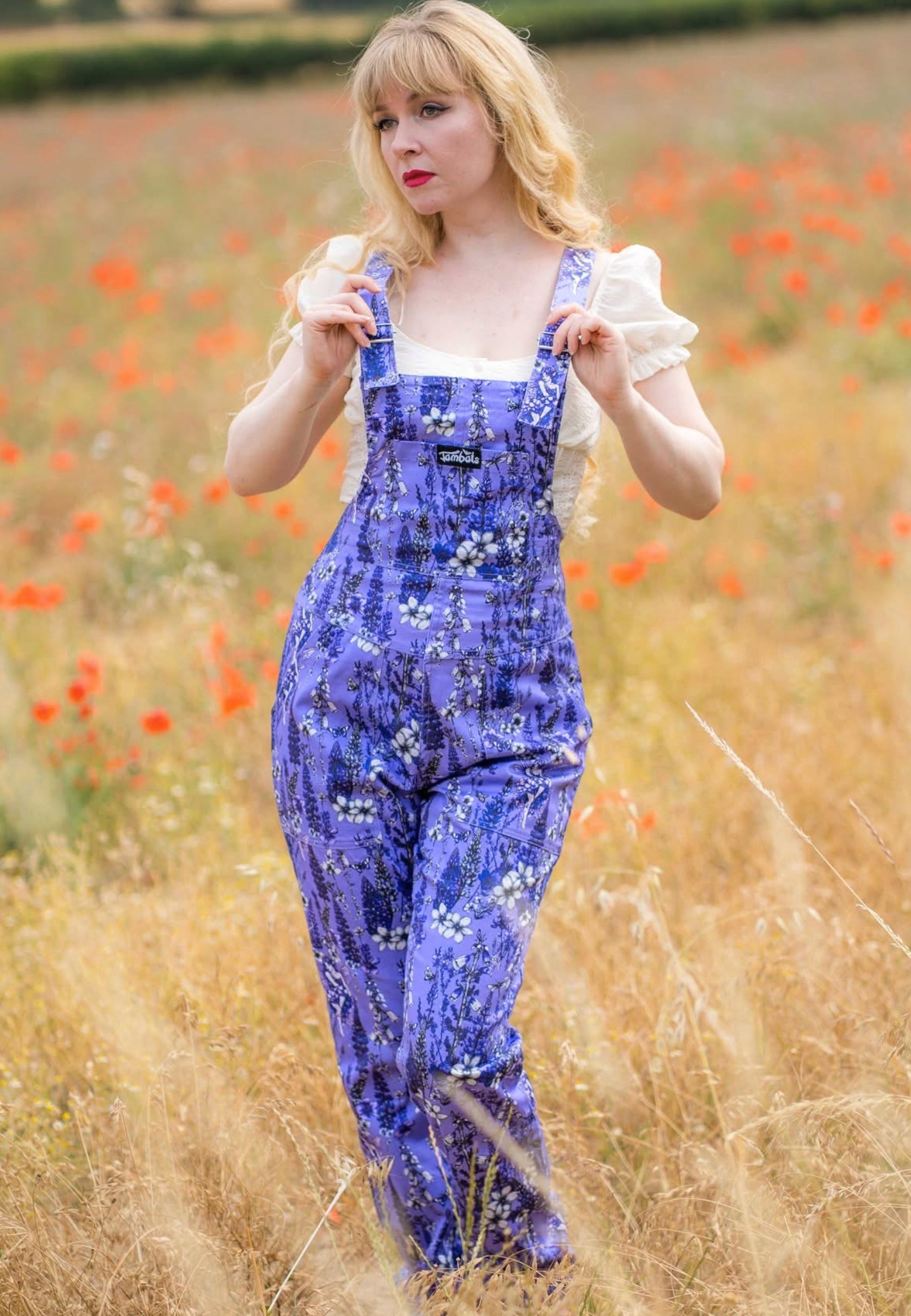 A woman standing in a field of red poppies, wearing a lilac overall dungarees with a white ruffled top. The dungarees has a beautiful whimsical print of wild flowers and flower fae 