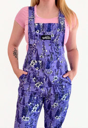 A model wearing purple floral print dungarees overalls over a pink t-shirt, shown from the neck down against a white background. The overalls feature a beautiful floral pattern of lavender, white flowers, and blue accents and delicate fae, with a black brand patch on the chest that reads 'Jambats'.