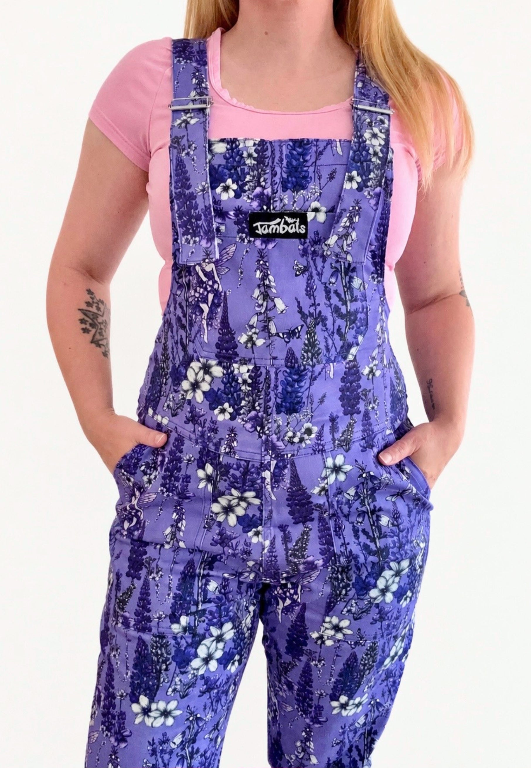 A model wearing purple floral print dungarees overalls over a pink t-shirt, shown from the neck down against a white background. The overalls feature a beautiful floral pattern of lavender, white flowers, and blue accents and delicate fae, with a black brand patch on the chest that reads 'Jambats'.