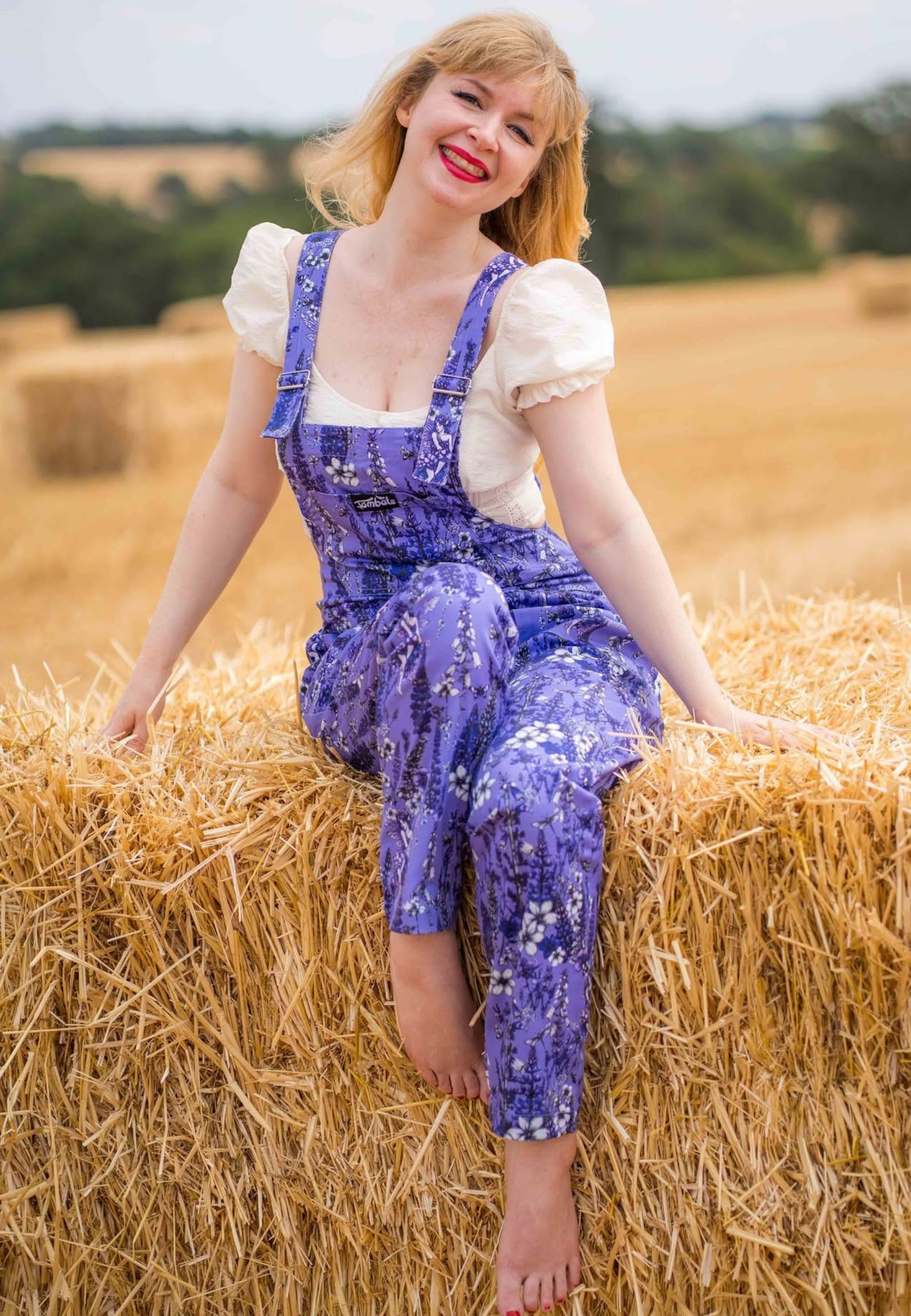 A woman sitting on a hay bale in a field, wearing a purple floral print overall dungarees over a white puff sleeve top.