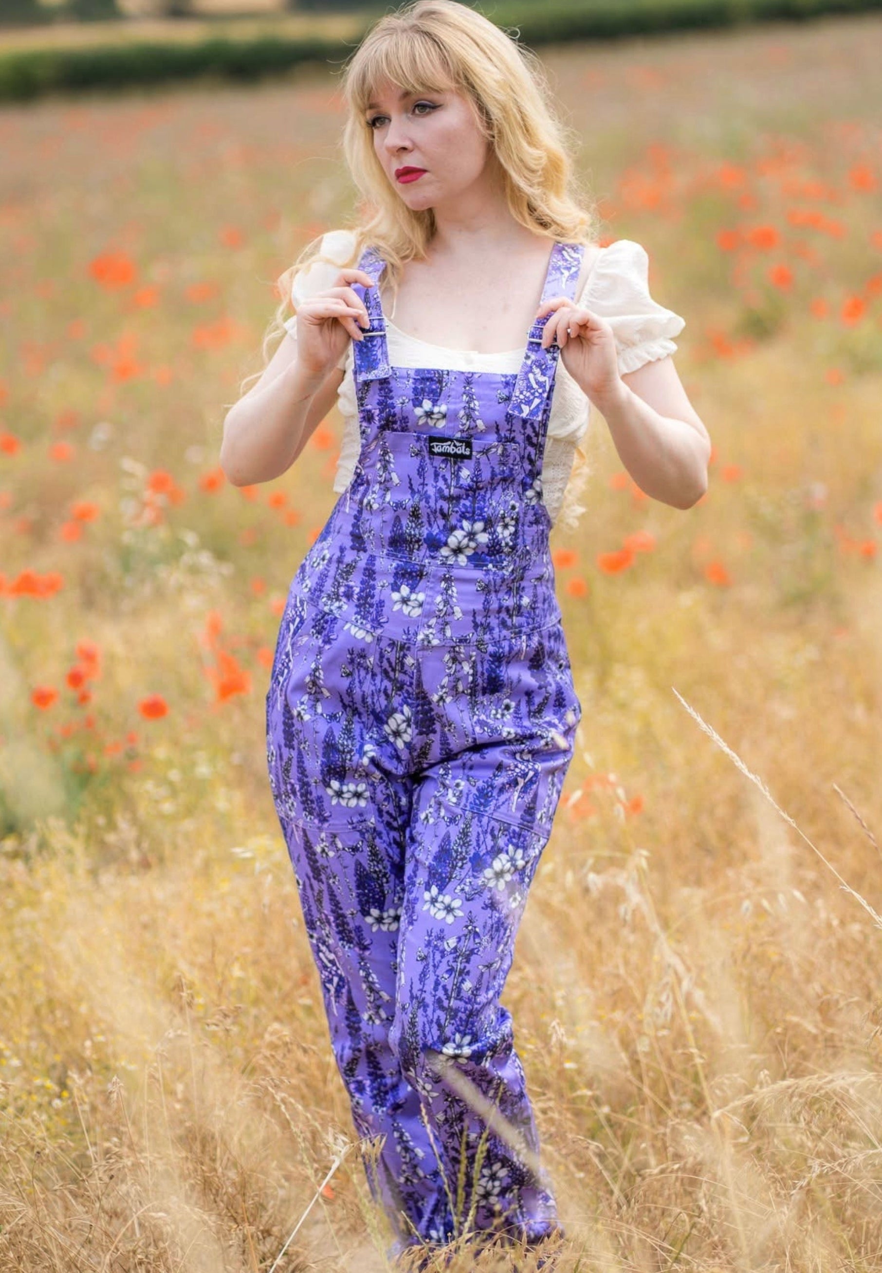 A woman standing in a field of red poppies, wearing a lilac overall dungarees with a white ruffled top. The dungarees has a beautiful whimsical print of wild flowers and flower fae 