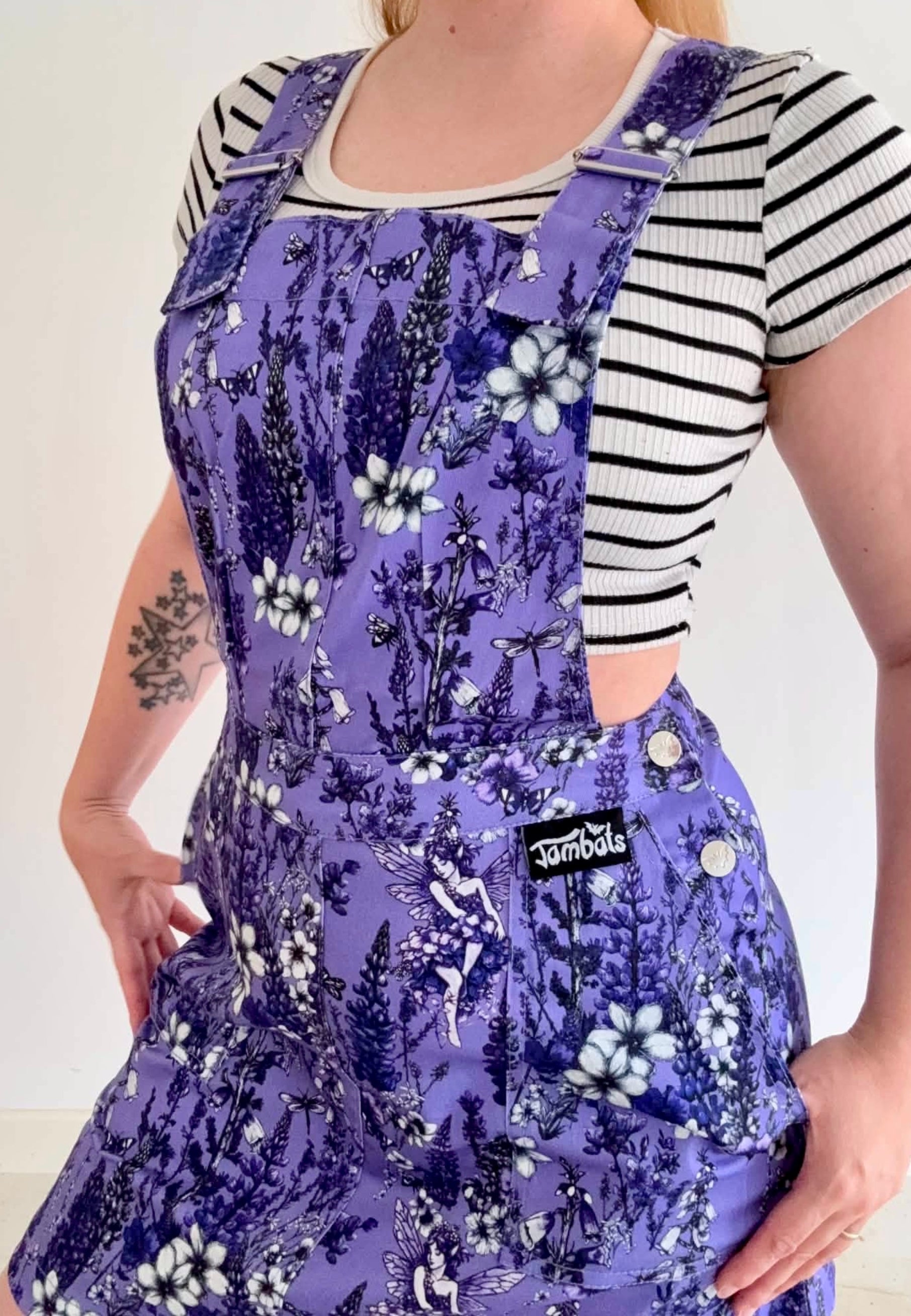 A woman wearing a deep lavender pinafore with a black and white striped t-shirt underneath. The pinafore features beautiful wild flowers and a Flower Fae, a front pocket, and a visible 'Jambats' brand patch on the side waistband 