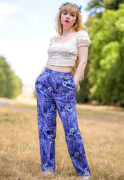 A woman standing outdoors in a grassy field wearing a cream puff-sleeve crop top and purple floral trousers, with a blue flower crown in her hair