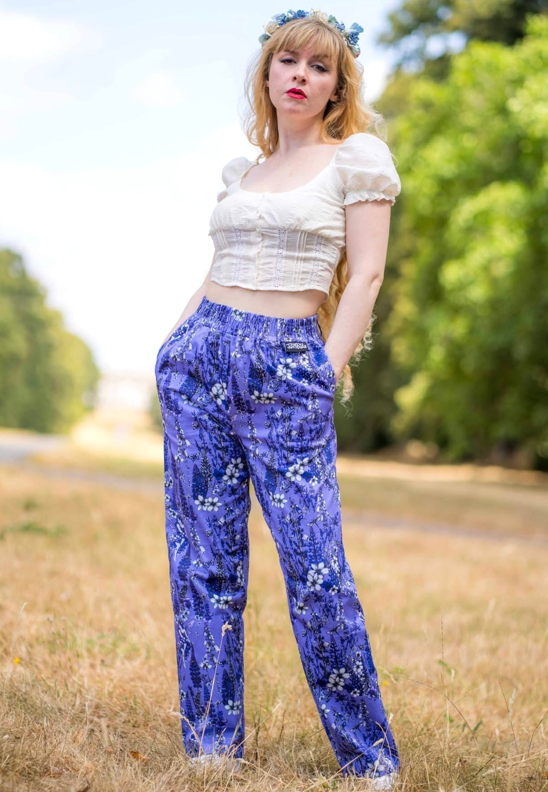 A woman standing outdoors in a grassy field wearing a cream puff-sleeve crop top and purple floral trousers, with a blue flower crown in her hair