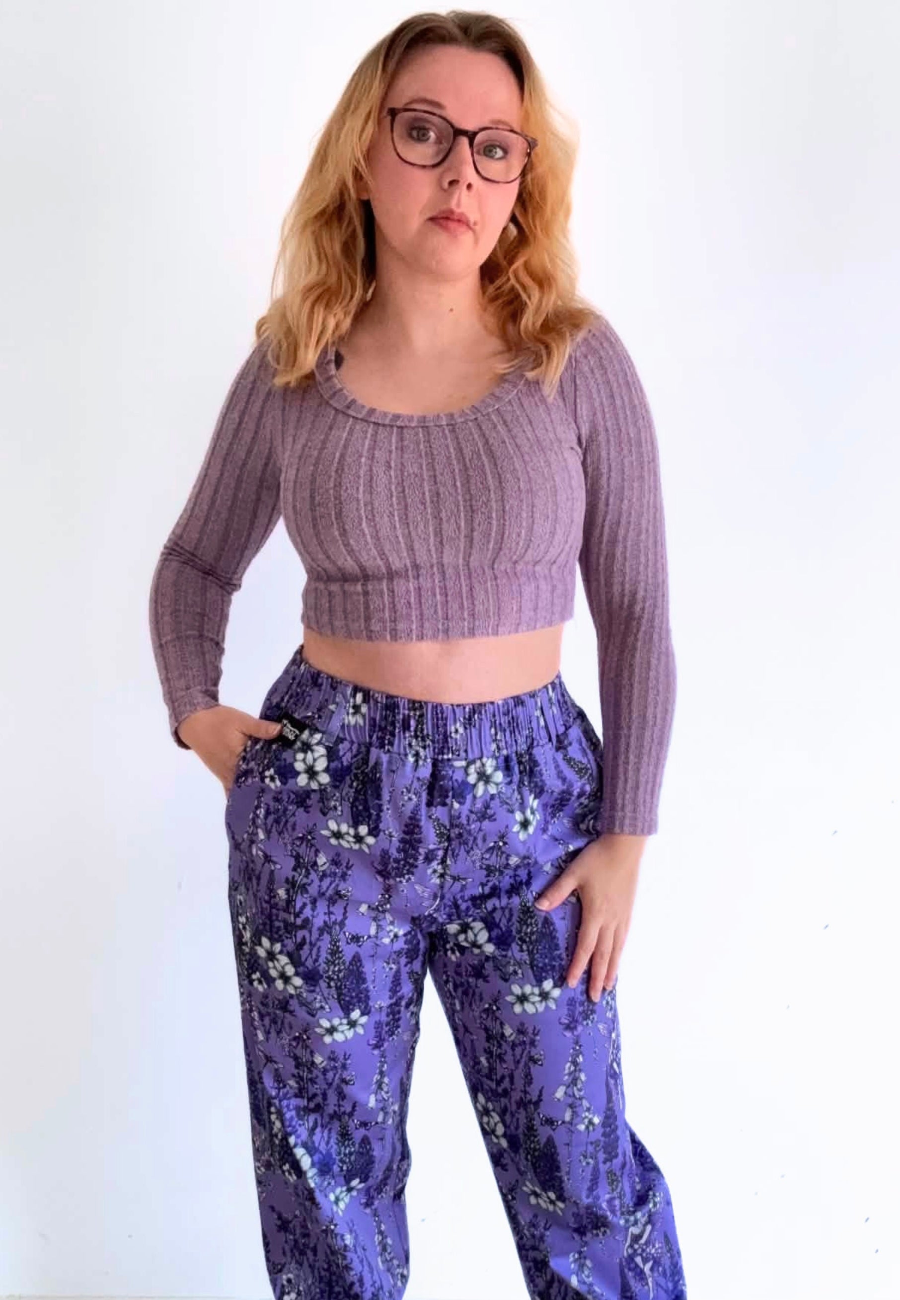 A woman wearing beautiful deep lavender colours trousers with a wild flower and fae print. Also wearing a dusky pink lilac crop top and glasses. Stood against a plain wall 
