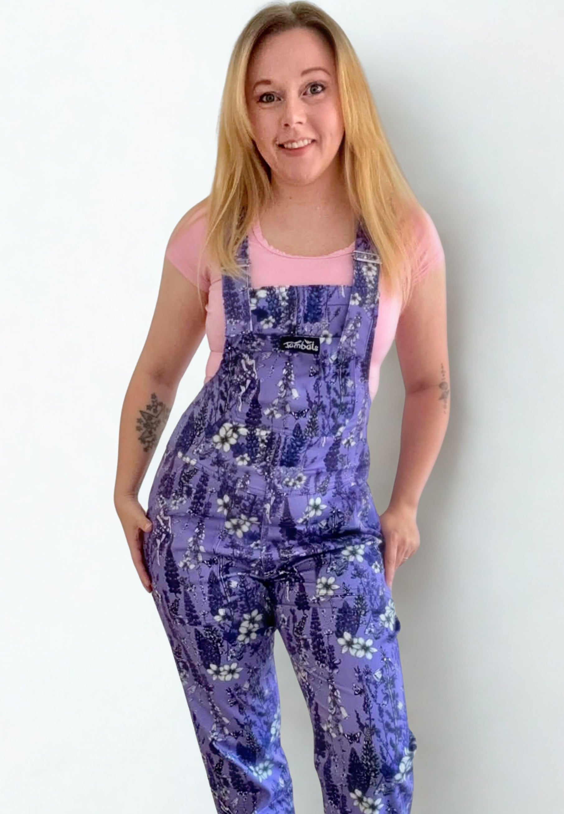 A woman modeling a pair of deep lavender floral print dungarees overalls over a light pink t-shirt, standing against a plain white background.