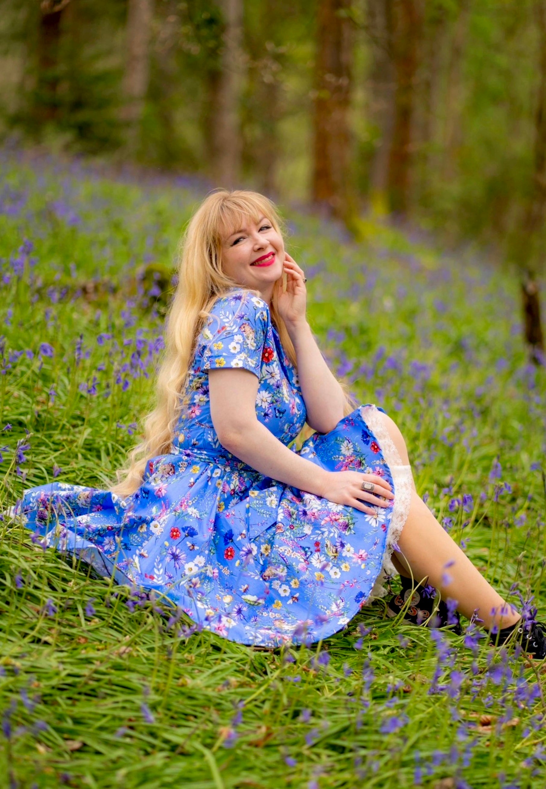Woman with very long blonde hair in a blue floral dress sitting in a field of bluebell flowers. Cottagecore romantic vibes. 