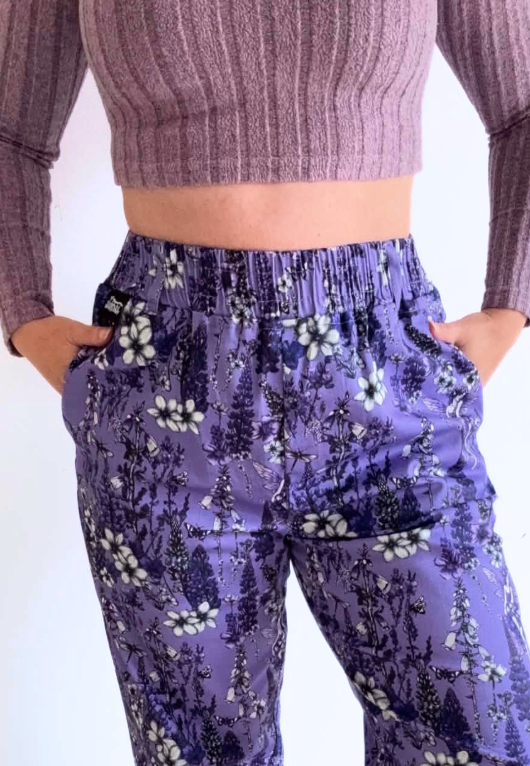 A person wearing lavender trousers with a beautiful illustrated print of wild flowers and fae. The model is also wearing a dusky pink purple ribbed long-sleeve crop top, standing against a plain white background.