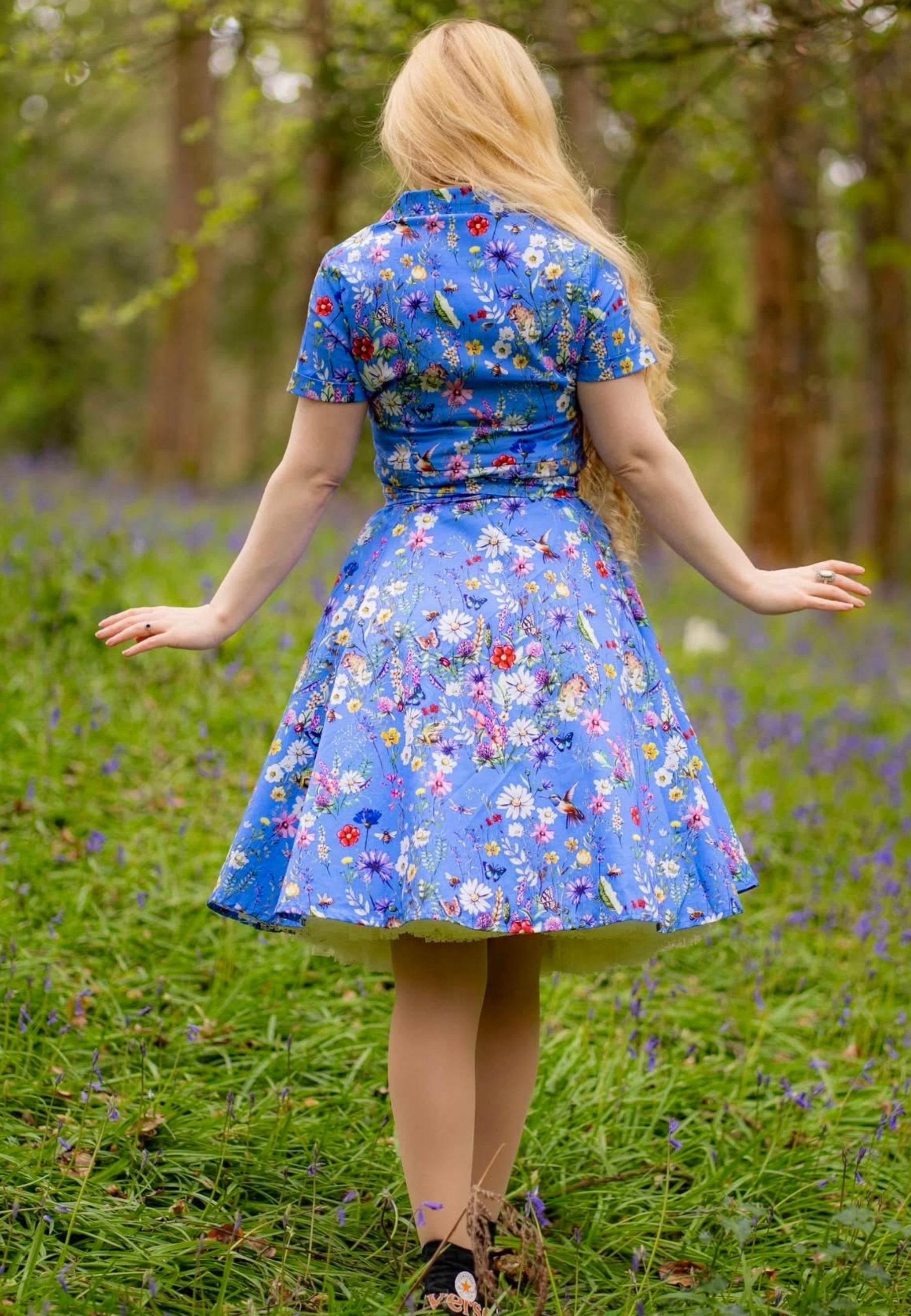 Woman with long blonde hair stood in a wild flower area of a wood wearing a beautiful cornflower blue dress with a wild flower print. Back view. 